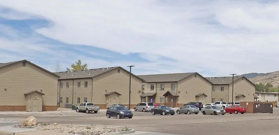 A row of beige two-story apartment buildings with small garages underneath, parked cars in front, power poles, and a mountain range in the background under a partly cloudy sky.