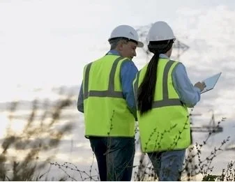 Two construction workers wearing white helmets and high-visibility vests are inspecting documents outdoors.