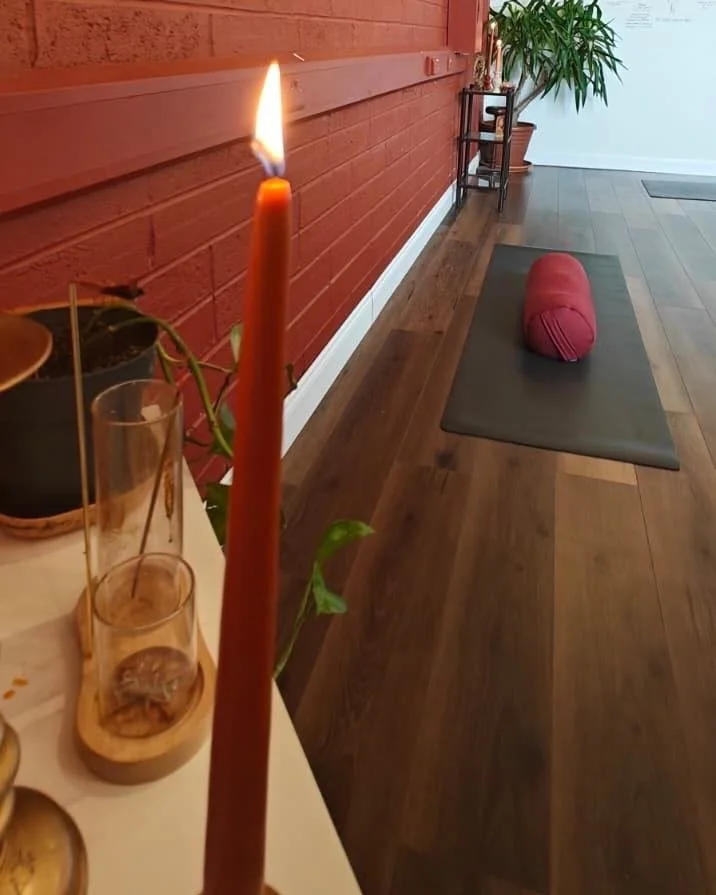 Lit red taper candle beside potted plants and glass holders in a yoga studio with a mat and bolster.
