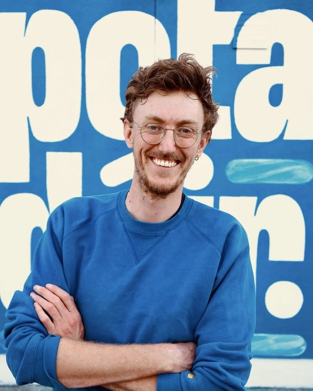 Smiling man wearing glasses and a blue sweatshirt, standing with crossed arms against a blue and cream graphic wall.