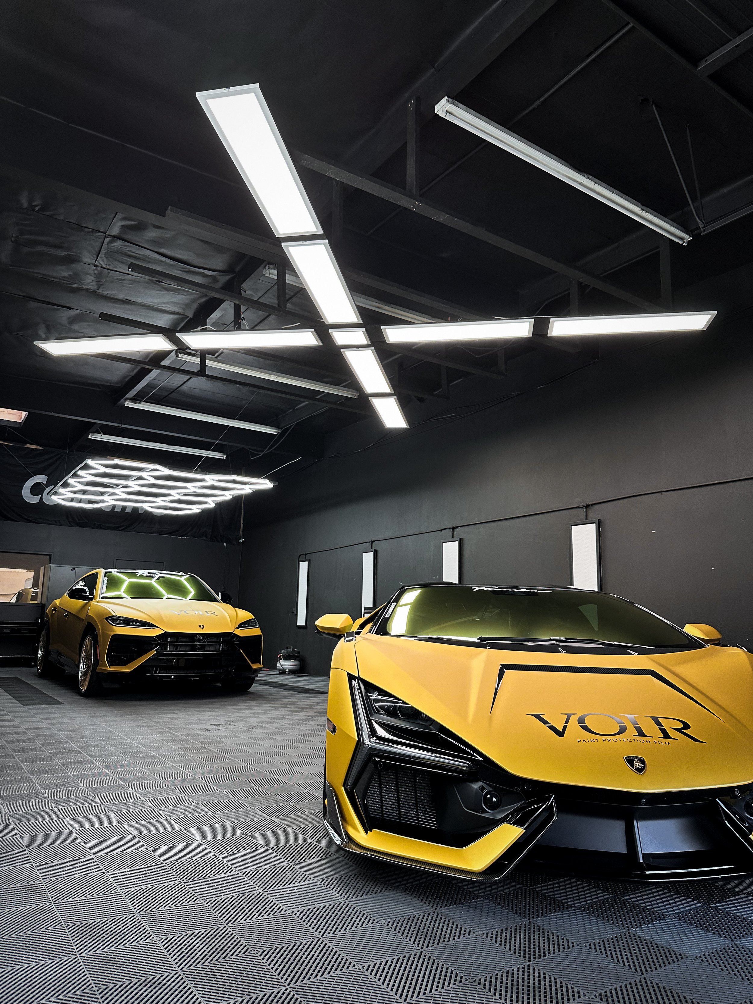 Two yellow luxury cars parked inside a modern showroom with black walls and unique white ceiling lights.