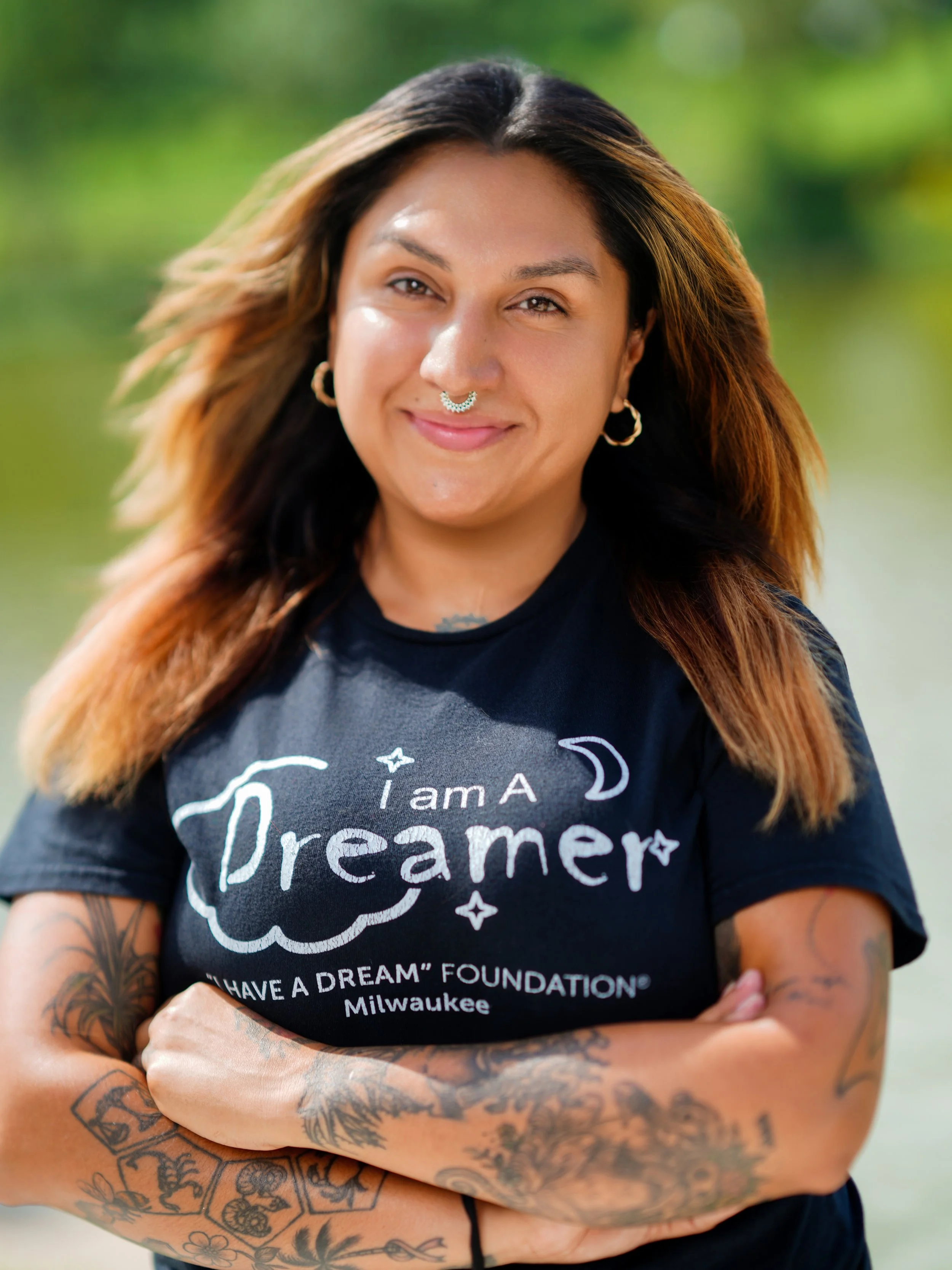 A woman with long, wavy hair and multiple tattoos wears a black t-shirt with white lettering that says 'I am a Dreamer' and 'Milwaukee'. She has a septum piercing, hoop earrings, and a slight smile. Her arms are crossed, and the background is blurred greenery.