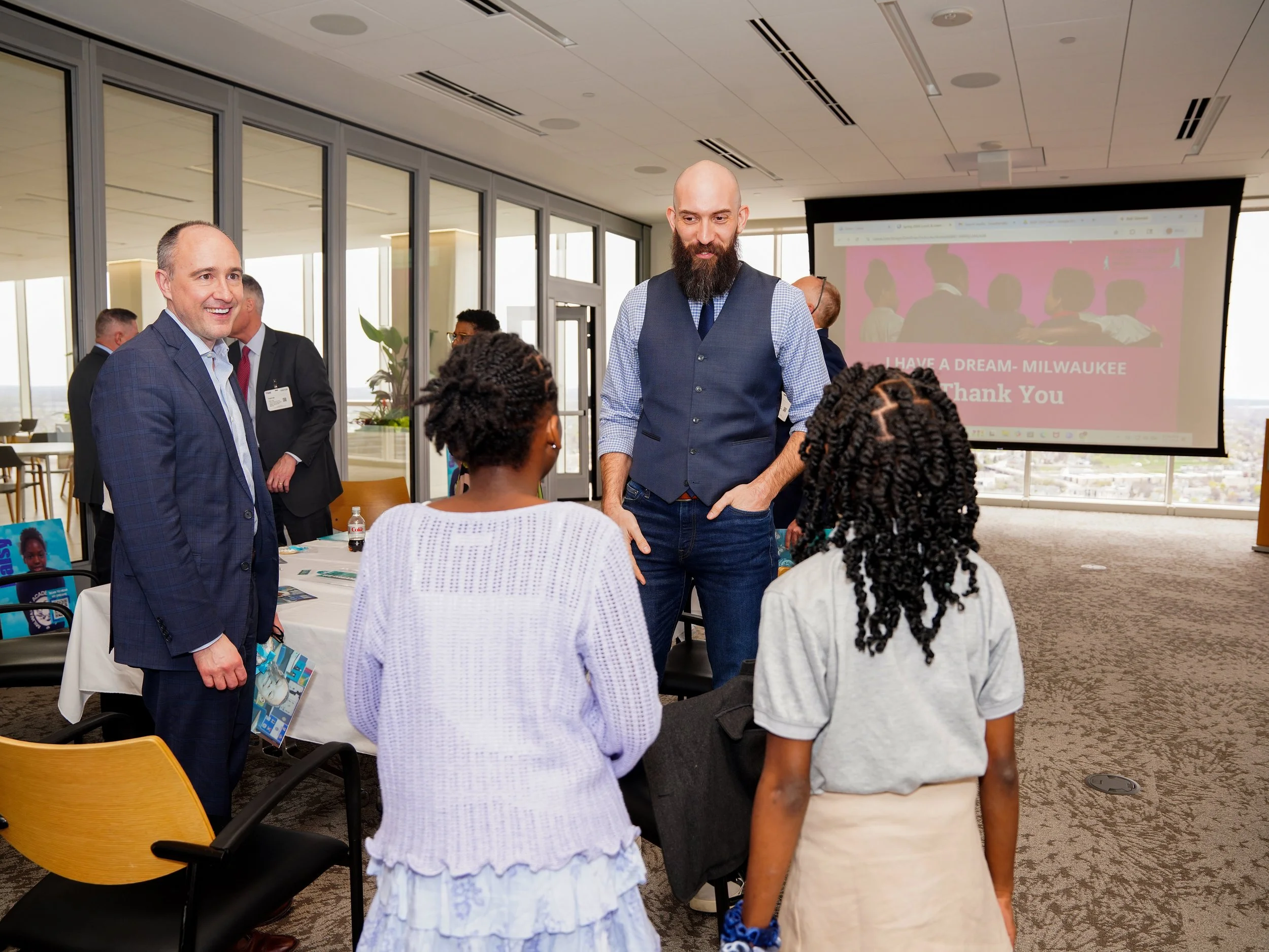 Group of people engaged in conversation at a professional event, with a presentation screen in the background displaying a message about a dream in Milwaukee.
