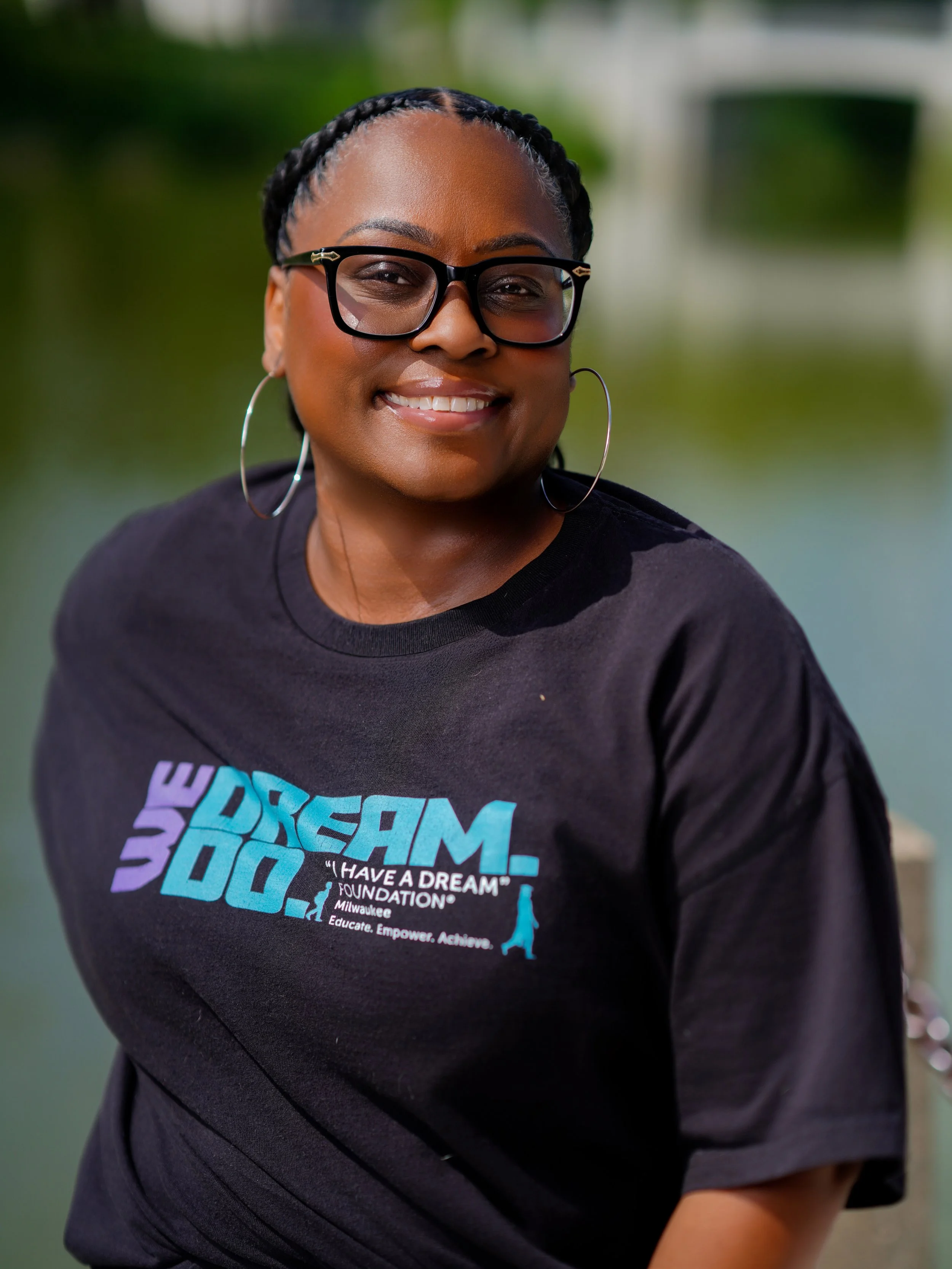 Close-up of a woman wearing glasses and hoop earrings, smiling outdoors with a blurred green background, wearing a black T-shirt with colorful text and graphics.