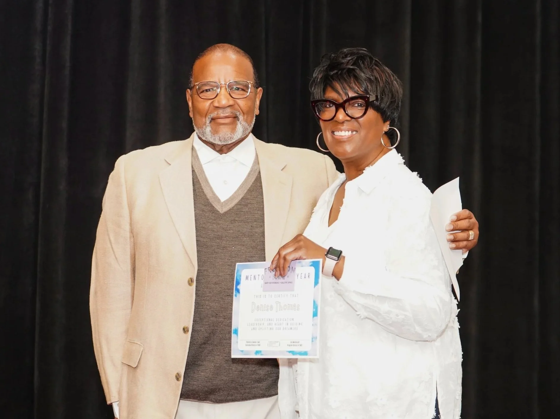 A man and woman standing together on stage, smiling. The man is wearing glasses, a beige blazer, and a light shirt. The woman has short dark hair, wearing large glasses, a white outfit, and is holding a certificate.