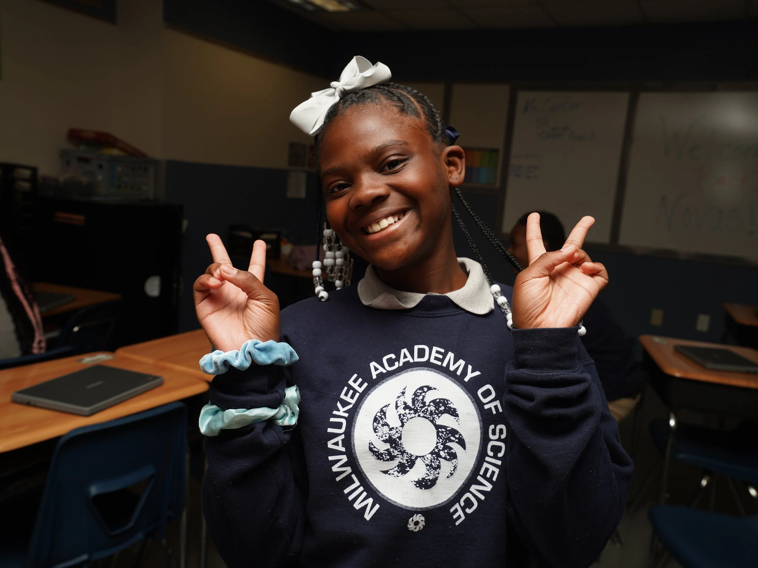 Smiling African American girl in school uniform making peace signs with both hands in classroom.