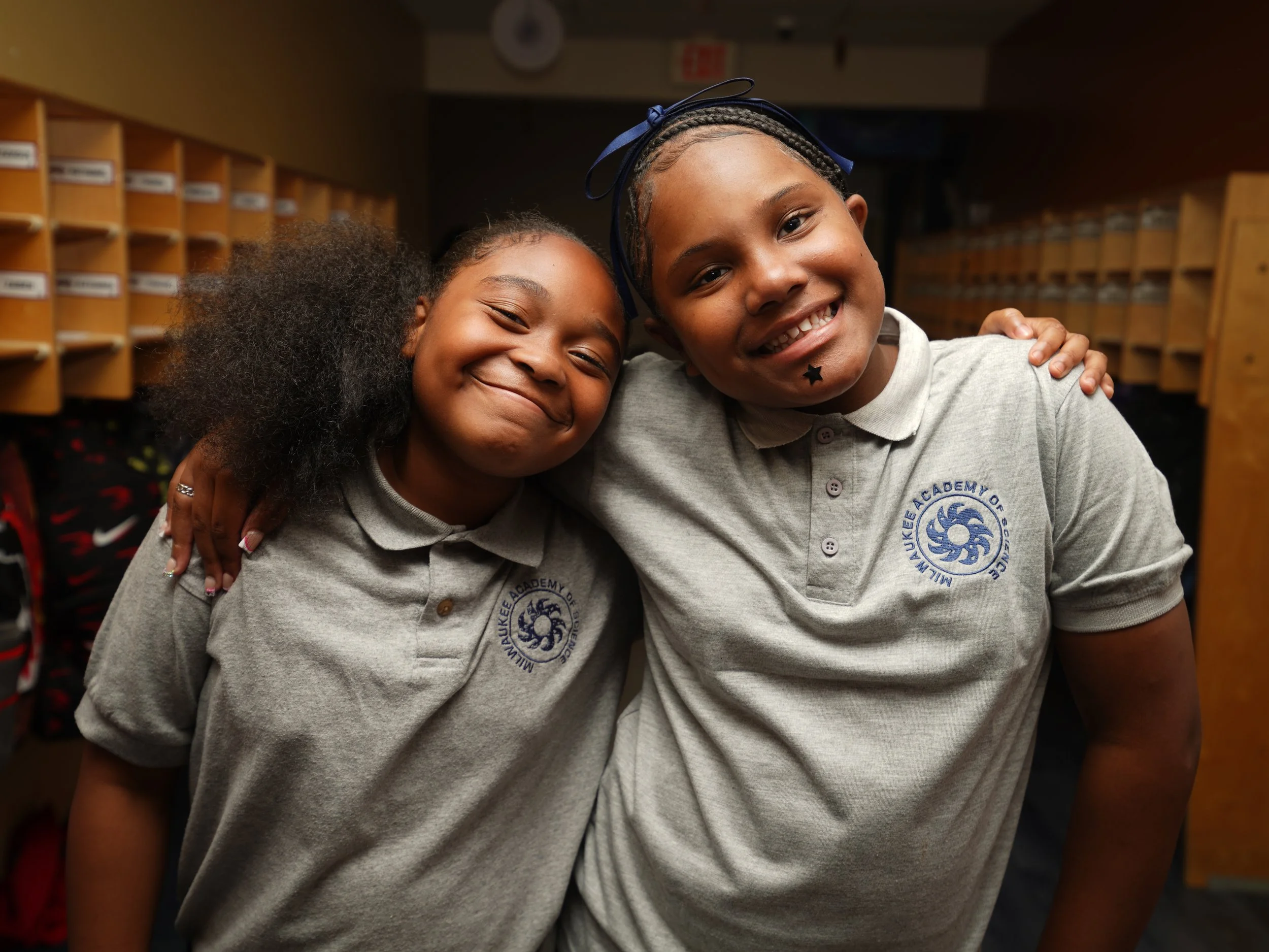 Two young girls smiling and hugging each other in a school hallway with cubby storage behind them.