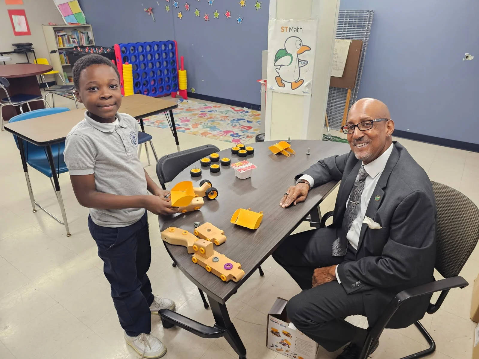 A young boy and an older man in a suit are sitting at a table with various yellow and black toy parts, assembling or playing with toy vehicles in a colorful classroom.
