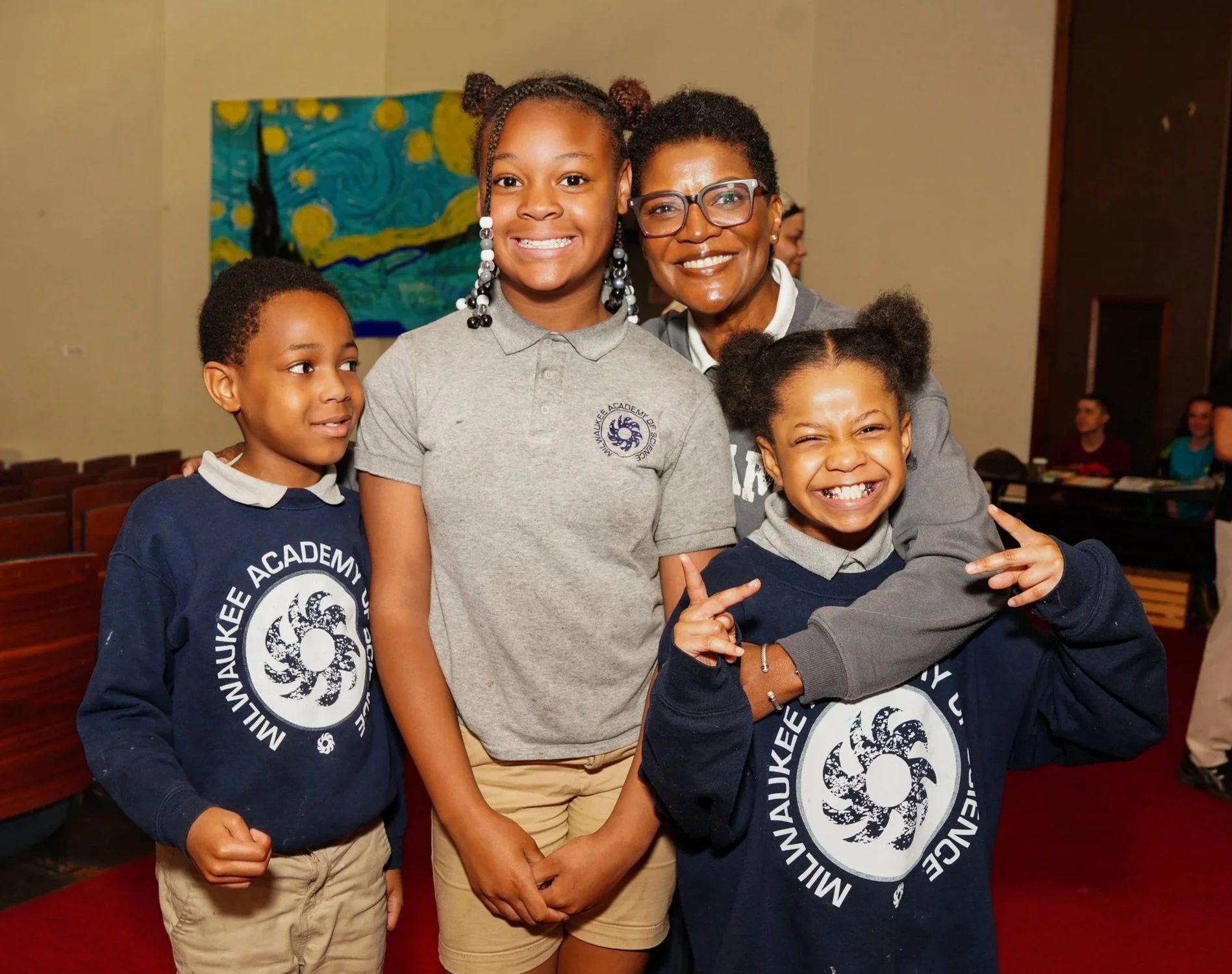 A woman and three children posing together indoors, smiling. The children are wearing navy blue sweatshirts with a circular logo, and the woman is dressed in a gray jacket and glasses. Art in the background.