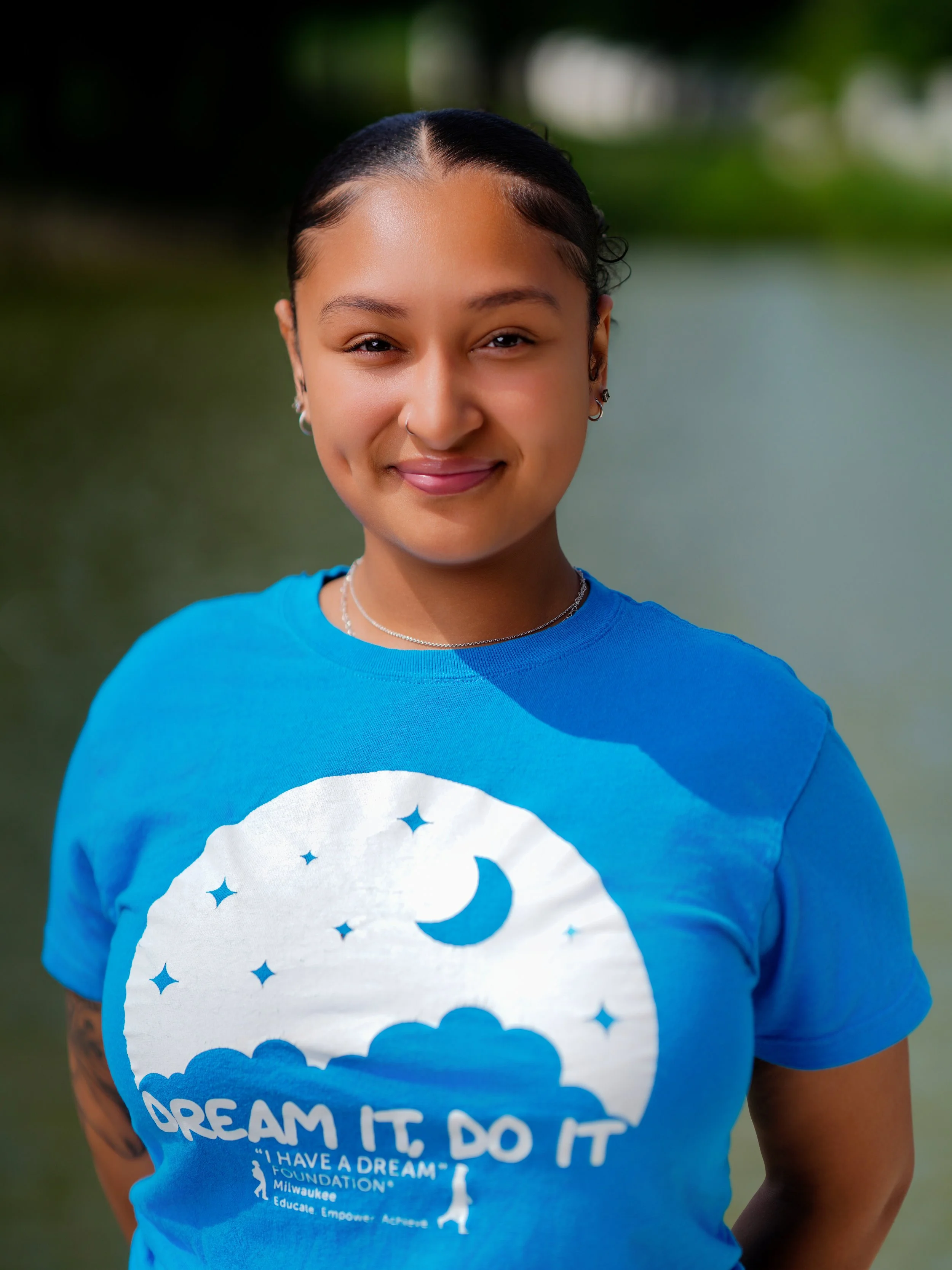 Close-up of a woman wearing a blue t-shirt with a white cloud and star design, smiling with eyes partially closed, with a blurred outdoor background.