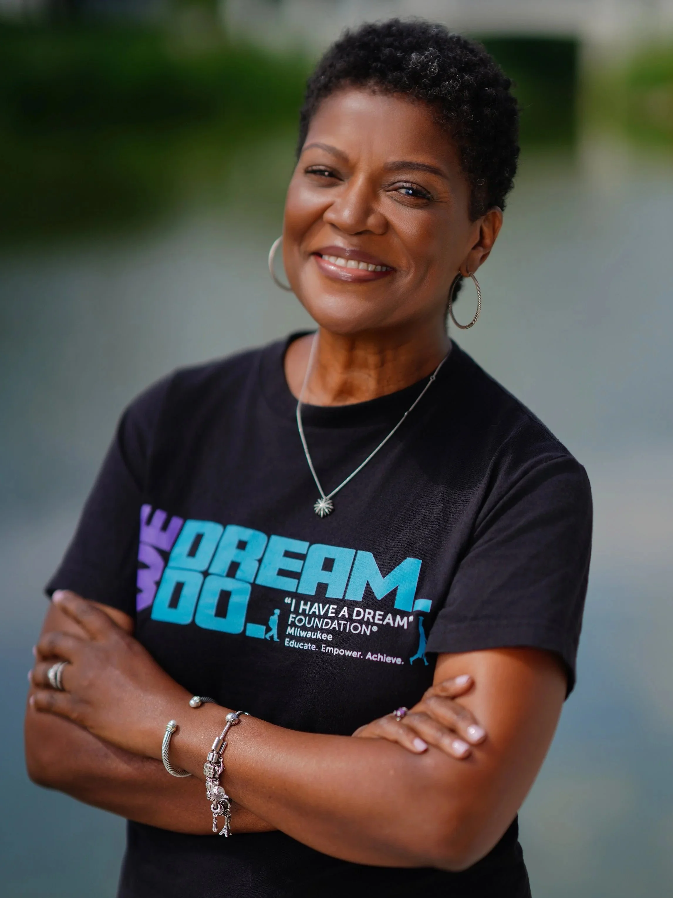 A woman with short curly hair, wearing hoop earrings, a necklace, and bracelets, standing outdoors with arms crossed. She is wearing a black T-shirt with colorful text and graphics, smiling and winking.