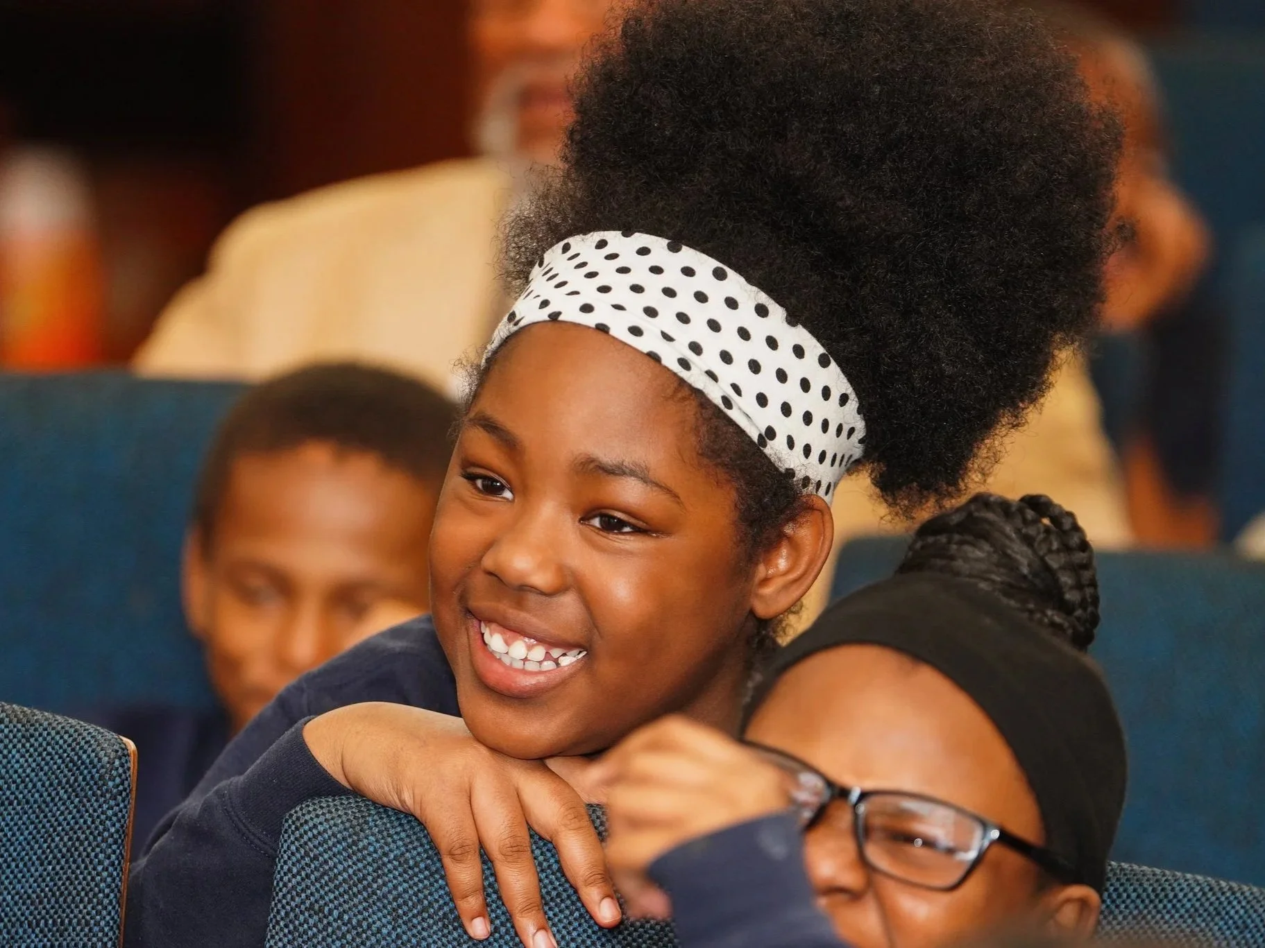 A young girl with a big smile, wearing a white polka dot headband and hugging someone in an indoor setting, with other people visible in the background.