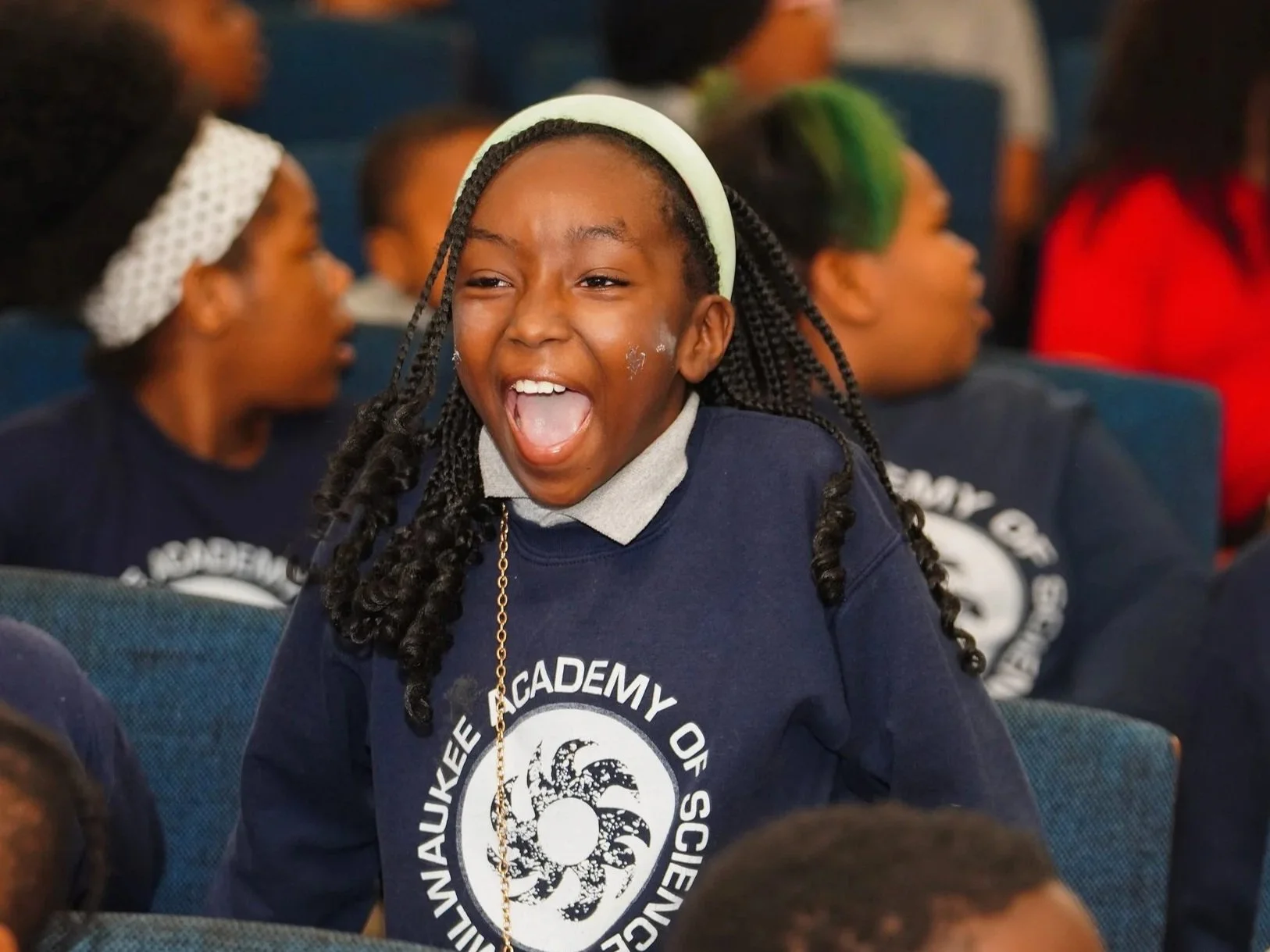 A group of children wearing navy blue shirts with the logo and name of a school or organization, sitting in an auditorium, with one girl with braids smiling and laughing in the foreground.