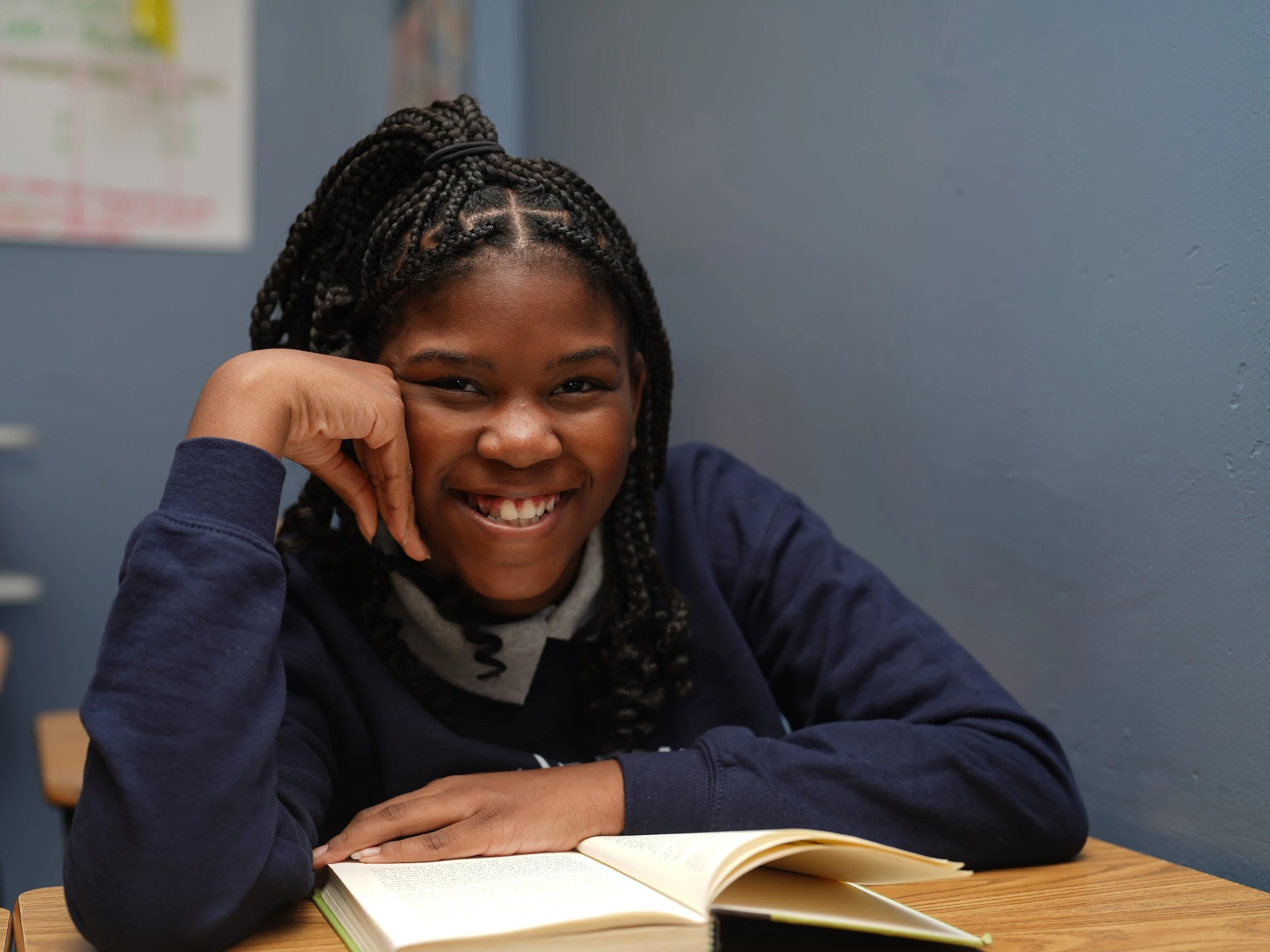 A young girl with dark braided hair smiling and resting her head on her hand while sitting at a wooden table with an open book in front of her. She is in a classroom with a blue wall and some posters in the background.