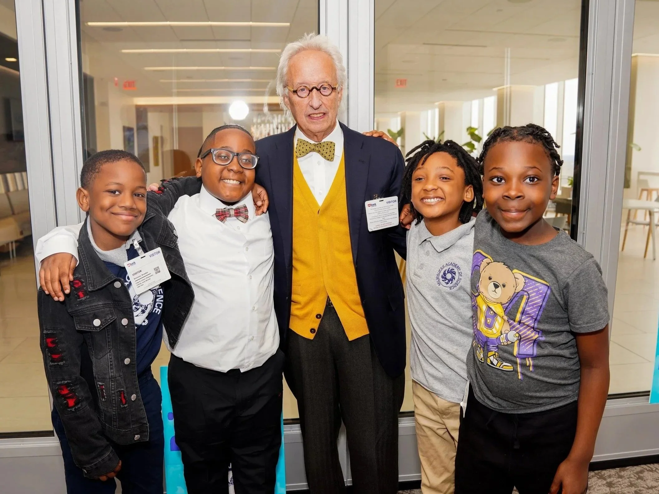 An elderly man with white hair, glasses, yellow sweater vest, and bow tie stands with four children inside a building. They are smiling and posing for the photo, with a glass door and modern interior in the background.