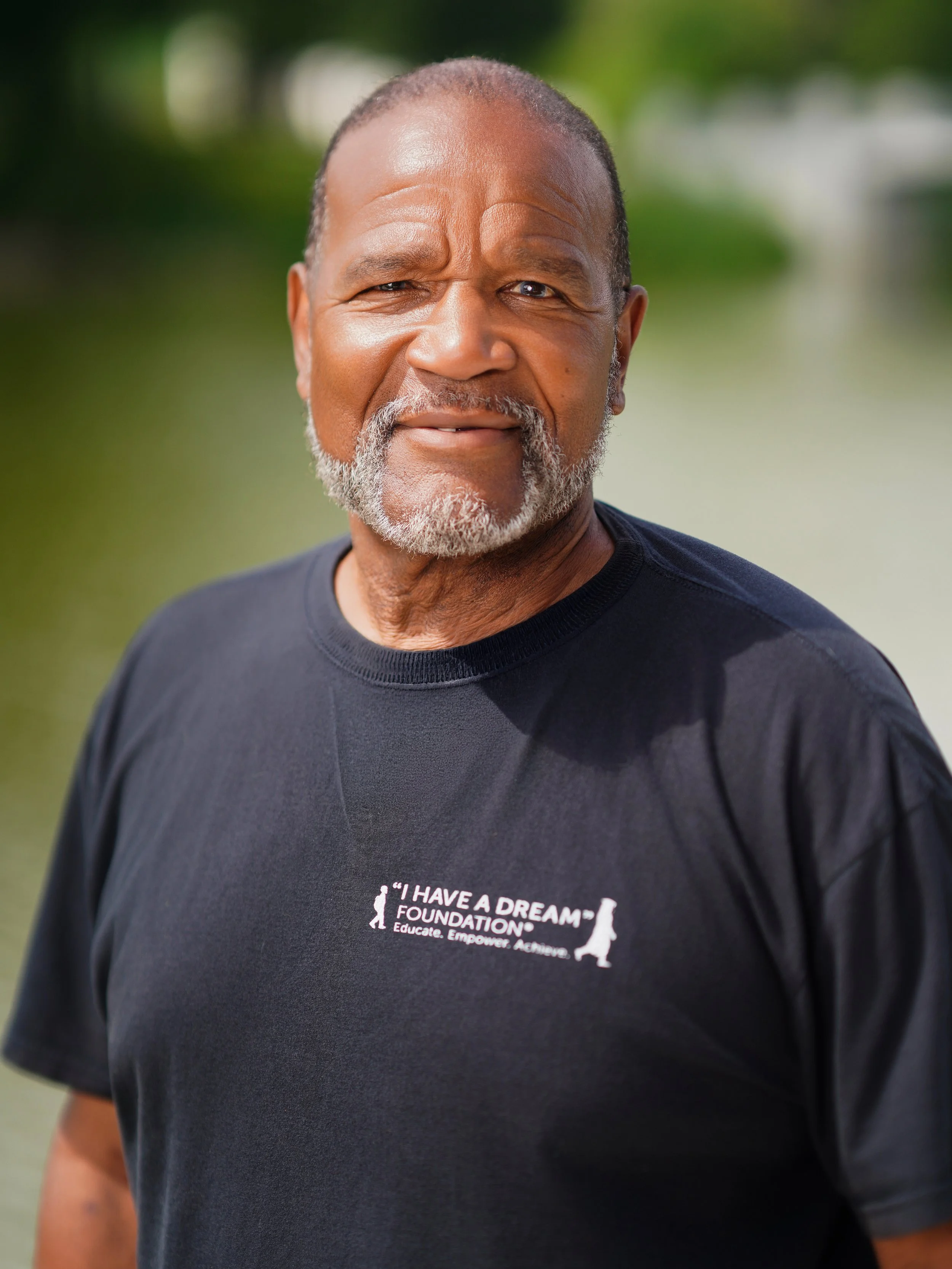 Close-up of a smiling elderly African American man wearing a black T-shirt with a logo and text that says 'I HAVE A DREAM FOUNDATION' with a small silhouette of a person walking and the words 'Educate. Empower. Achieve.' in front of a blurred green outdoor background.