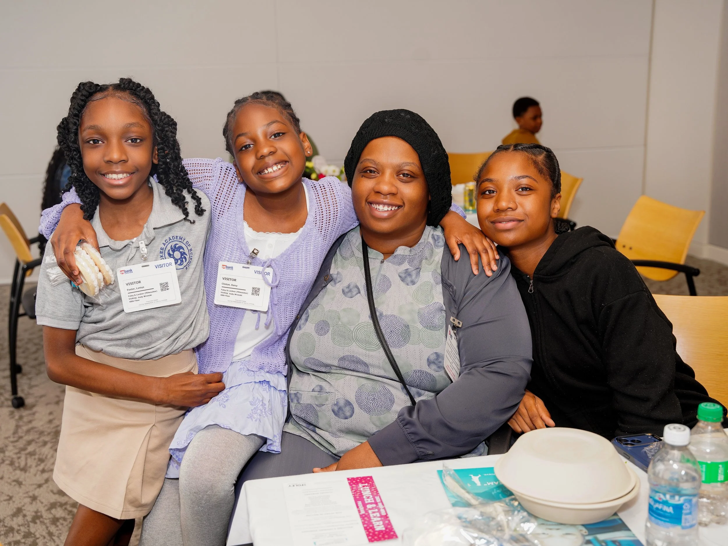 Four smiling African American girls sitting at a table, three with their arms around each other, in a room with chairs and a boy in the background. They wear casual clothing and have visitor badges around their necks.