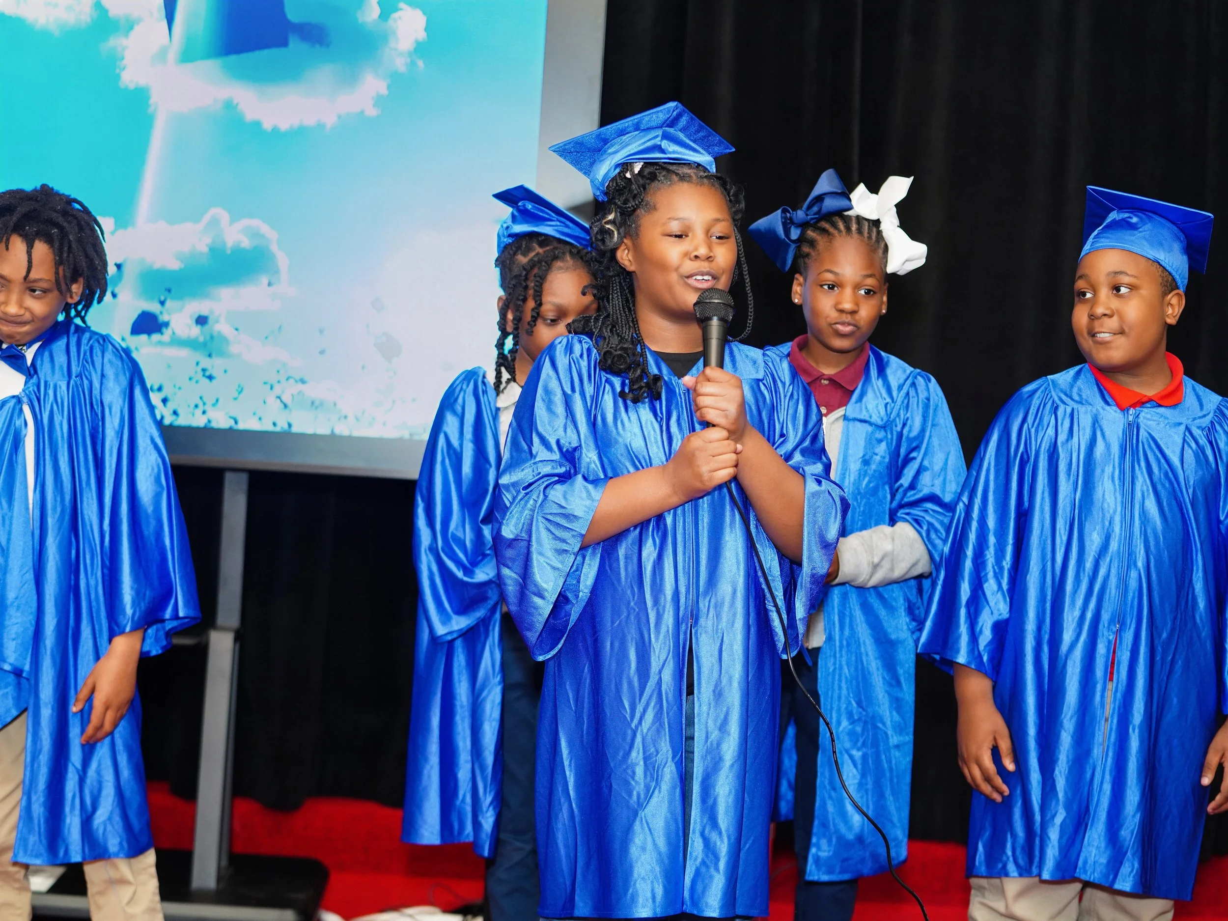 Young children in blue graduation gowns and caps on stage, one girl holding a microphone, during a graduation ceremony.