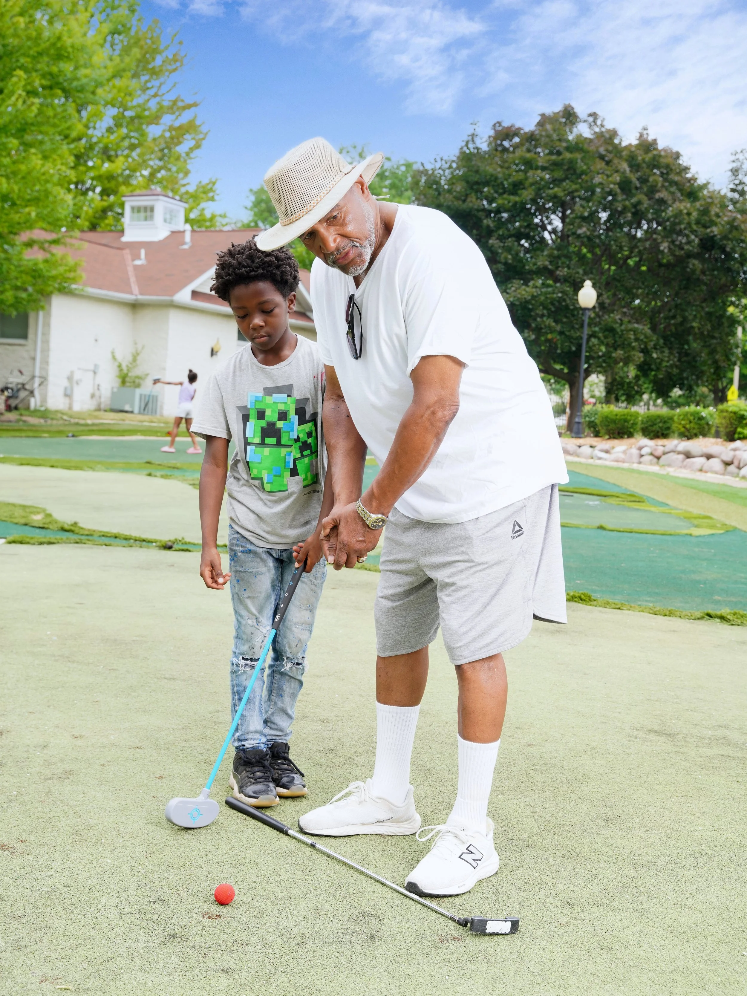 An older man and a boy playing mini golf outdoors on a sunny day. The man is wearing a light-colored hat, white t-shirt, and gray shorts, while the boy is dressed in a gray T-shirt with a video game character and distressed jeans. They are standing on a putting green with trees and a house in the background. The man and boy are focused on hitting a small red golf ball with a putter.