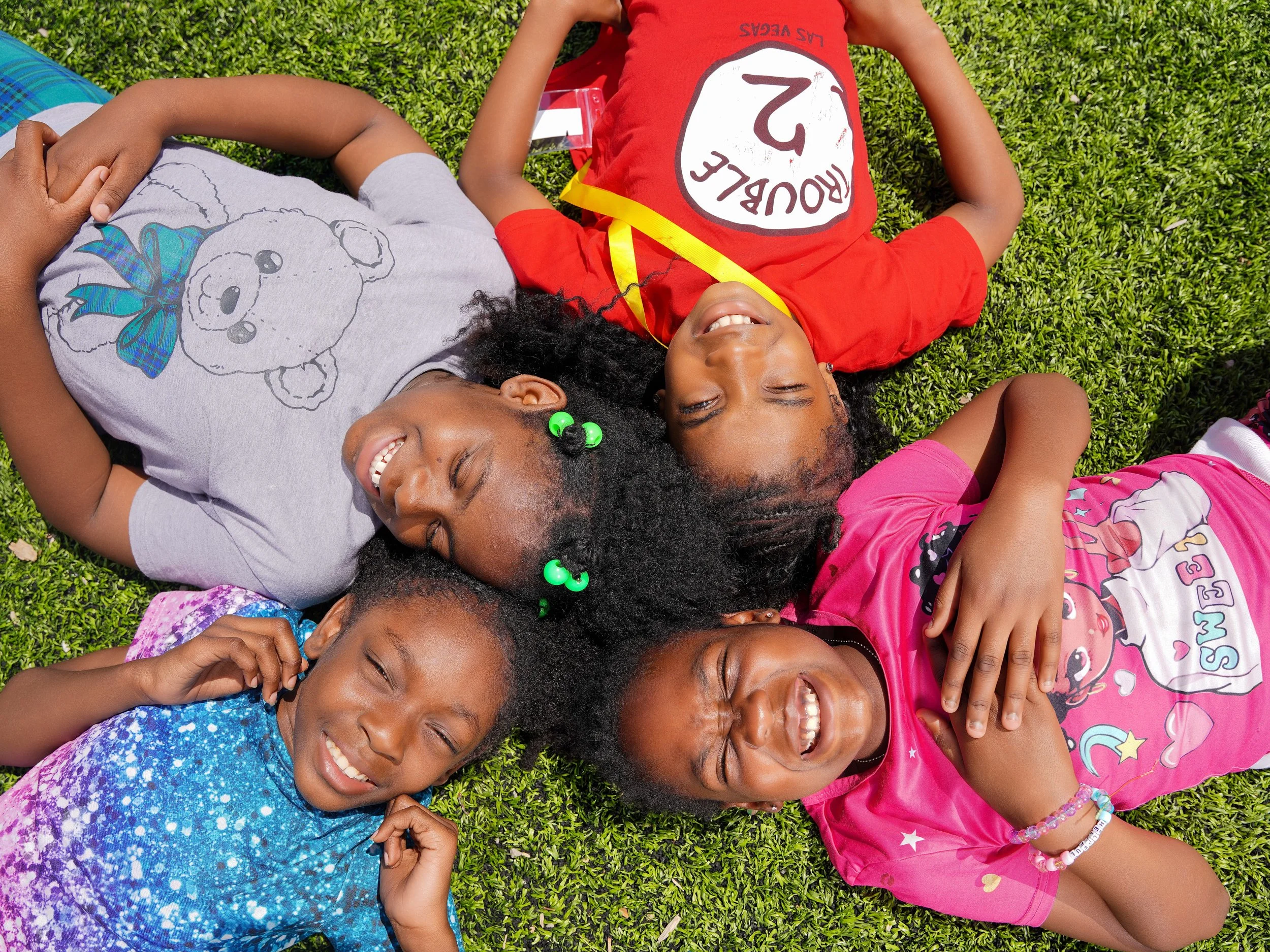 Group of children lying on grass, smiling and laughing, wearing colorful clothes.