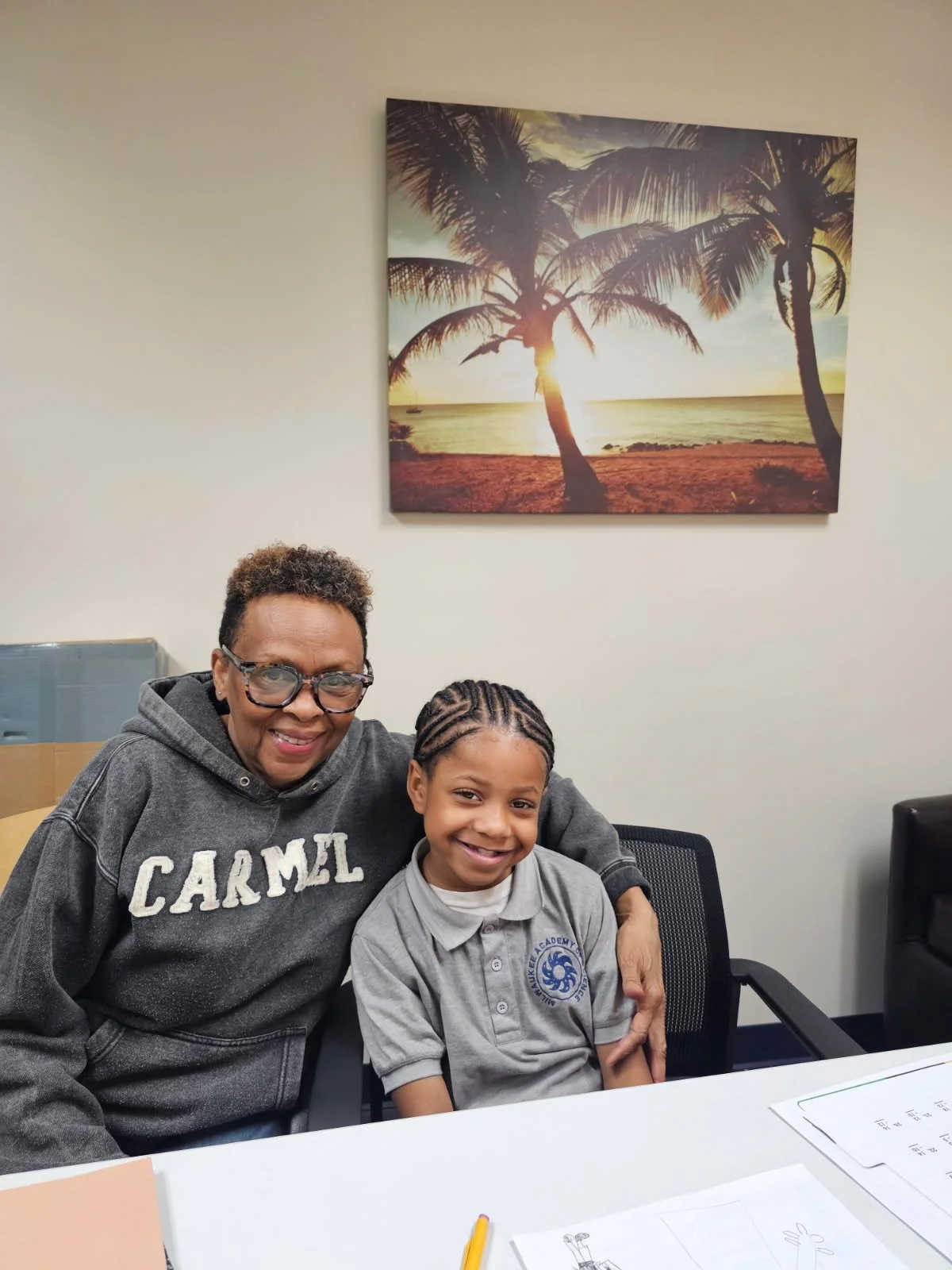 A woman and a young girl sitting together at a table in an office, smiling at the camera, with a sunset beach scene painting on the wall behind them.