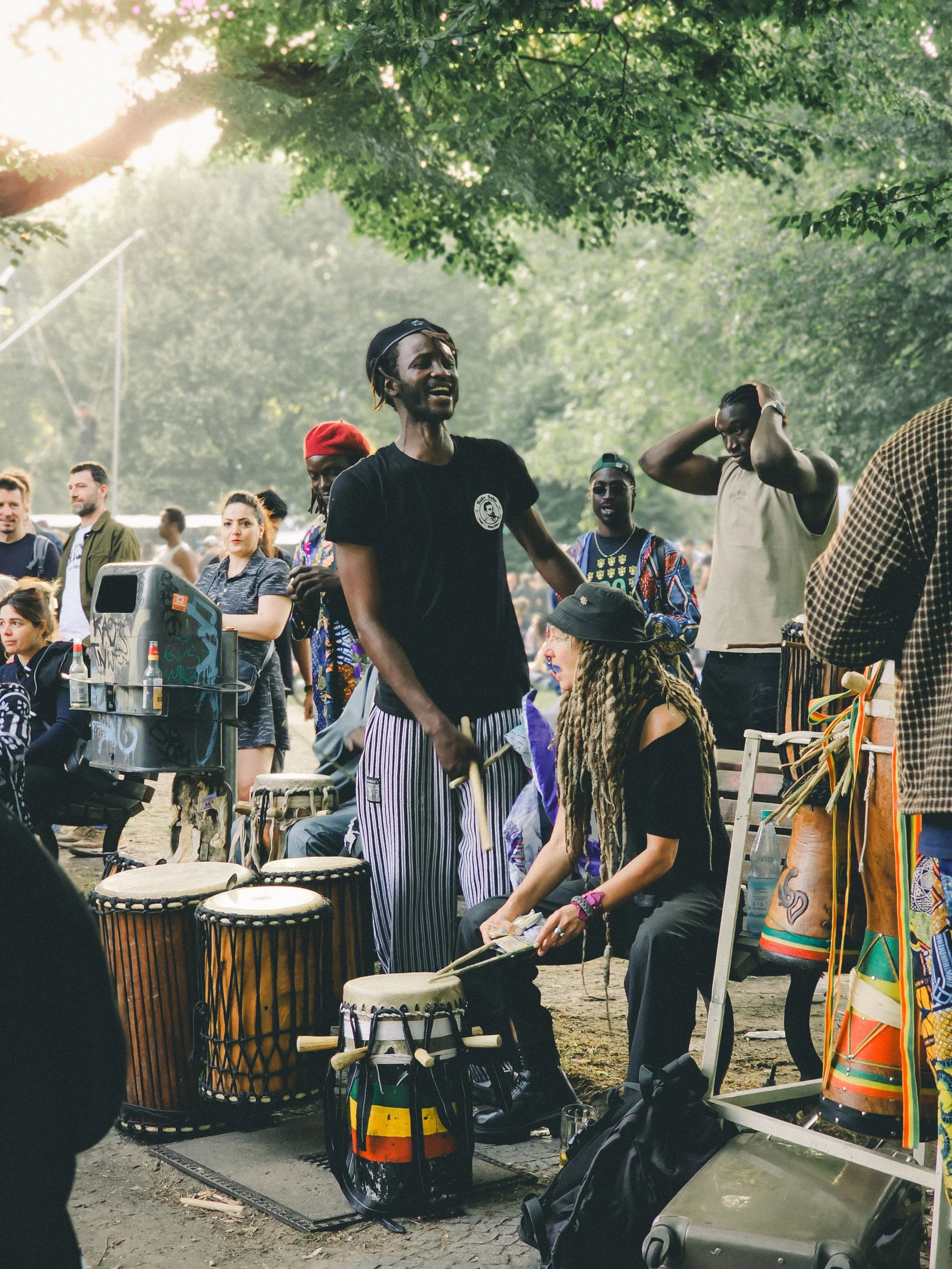 A diverse group of people, including African and Caucasian individuals, participating in drumming and musical performance outdoors under large trees with sunlight filtering through the leaves. Some are playing drums while others are watching or preparing to join in.