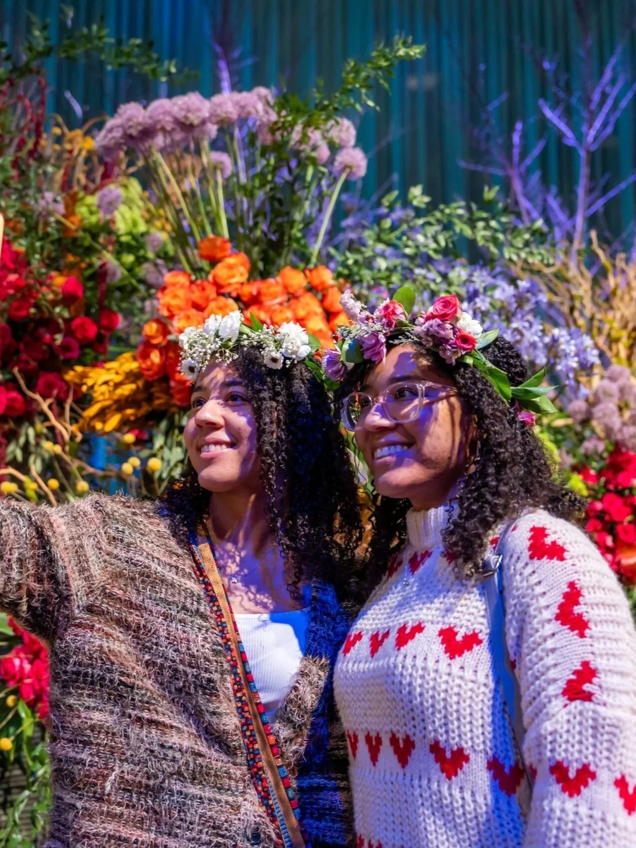 Two women with curly hair wearing flower crowns and colorful sweaters, taking a selfie in front of a vibrant floral backdrop.