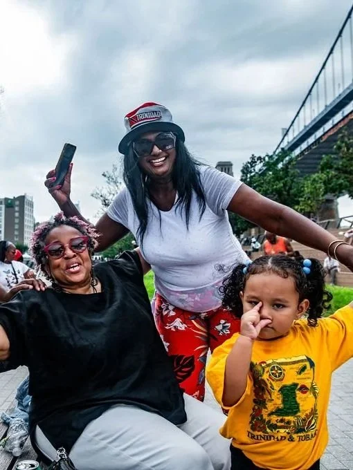 Three women posing outdoors, smiling and enjoying themselves. One woman is wearing sunglasses and a black outfit, another woman is wearing a white shirt, red patterned pants, a red and white Santa hat, and sunglasses, and the young girl is wearing a yellow shirt and has black curly hair.
