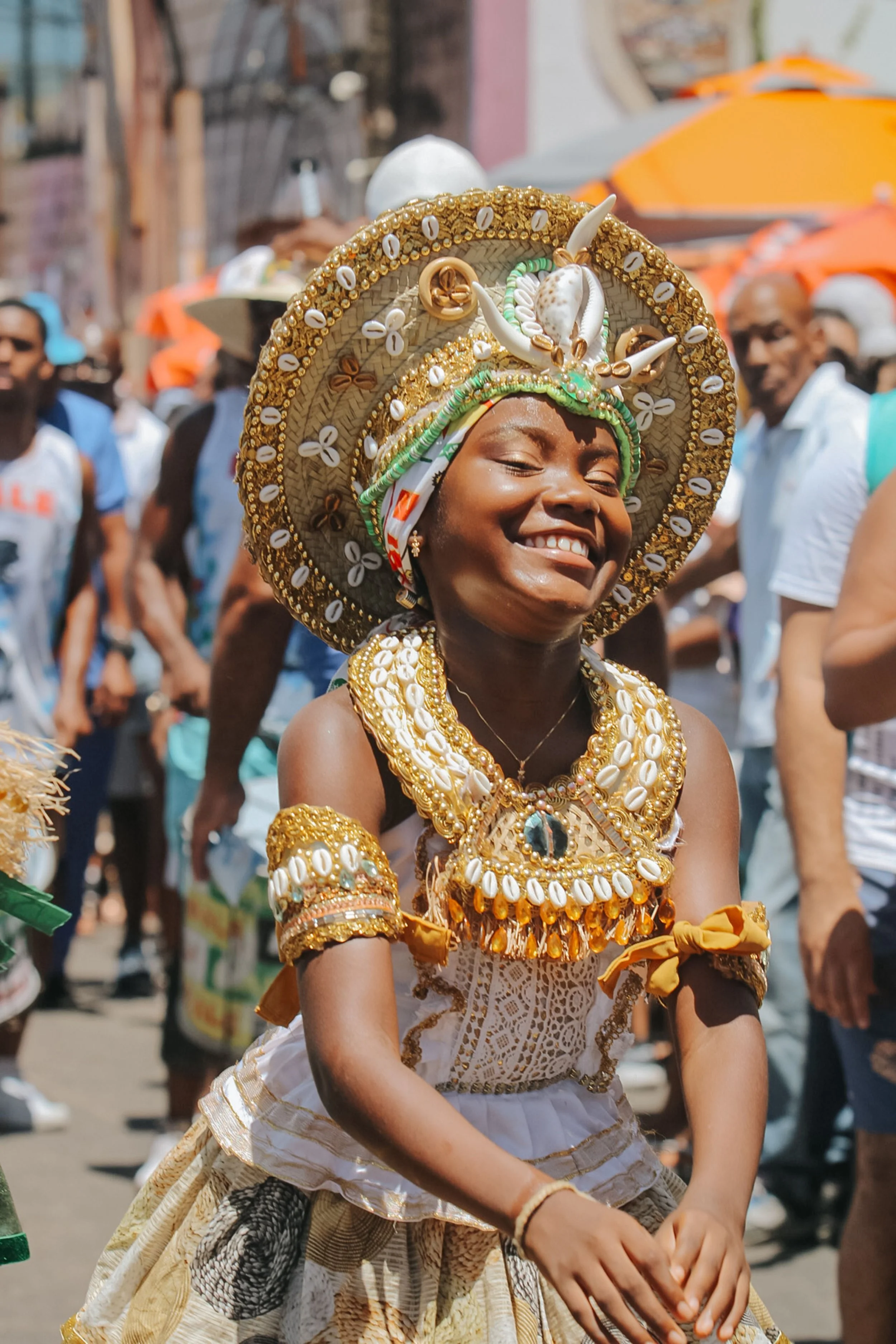 Young woman smiling and dressed in elaborate traditional costume with gold and shell adornments during a parade or celebration in an urban setting.