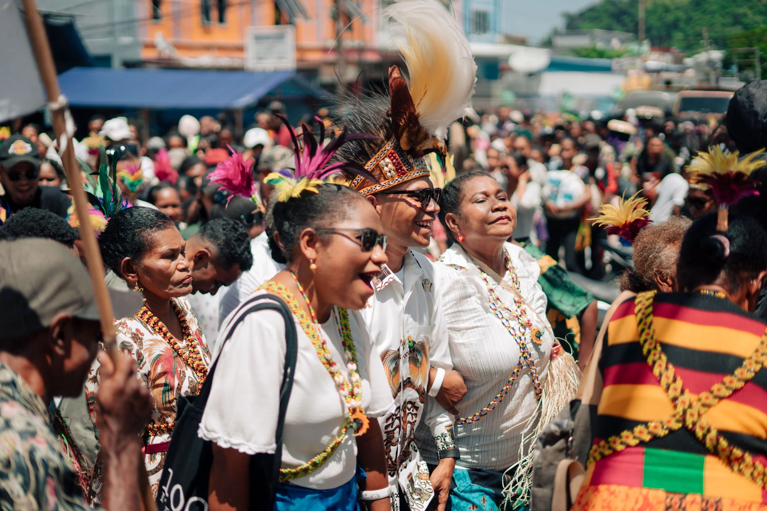 A group of people celebrating outdoors, wearing traditional colorful clothing and feathered headdresses, with some smiling and others looking pensive. The background shows a bustling street scene with buildings and many more people.
