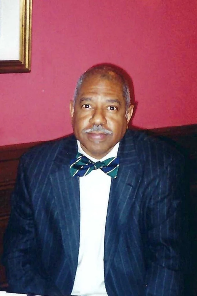 A man wearing a dark pinstripe suit and a green and blue striped bowtie, sitting against a red wall with a framed mirror to the left.