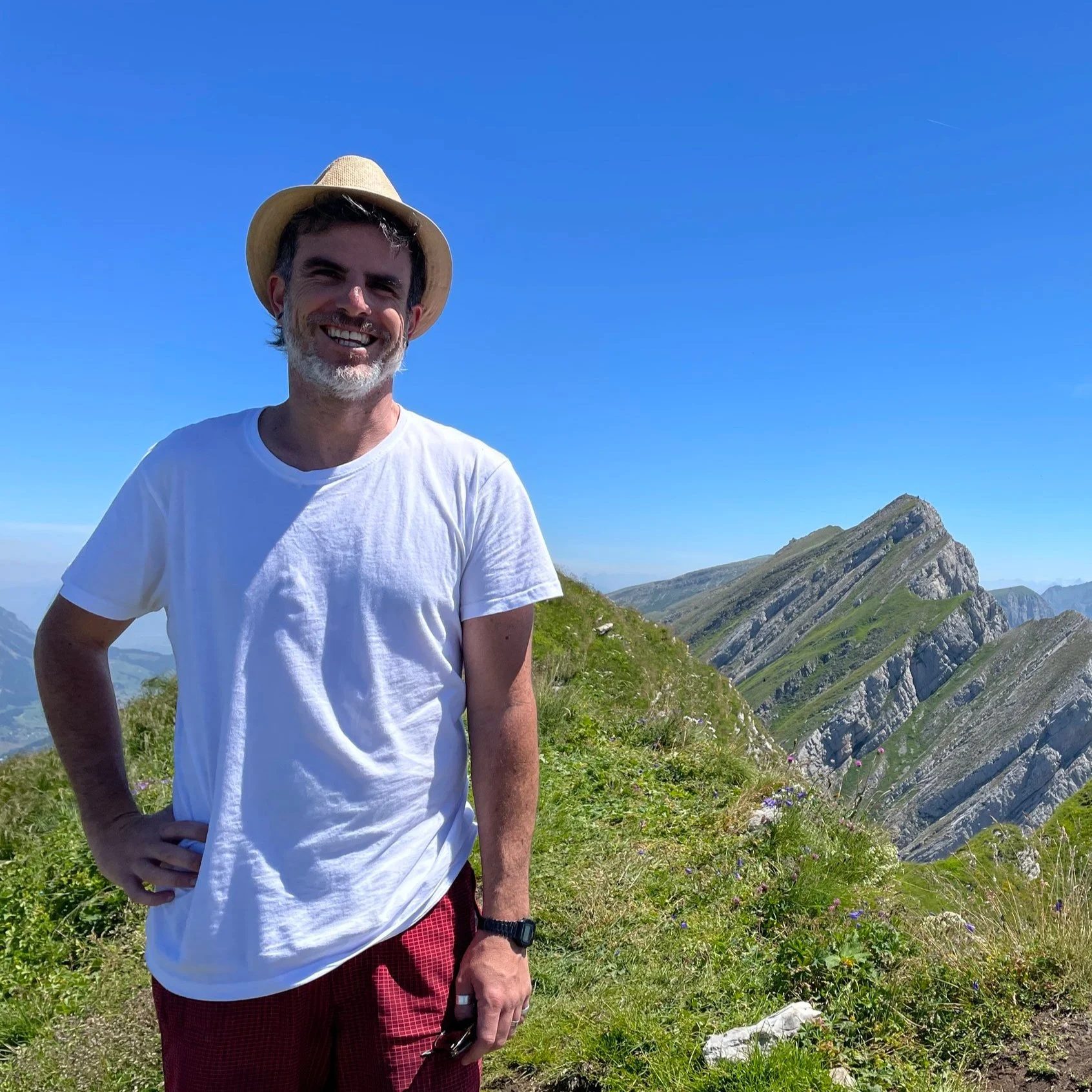 A smiling man wearing a white T-shirt, red shorts, a watch, and a straw hat standing on a grassy mountain trail with green hills and rocky peaks in the background under a clear blue sky.