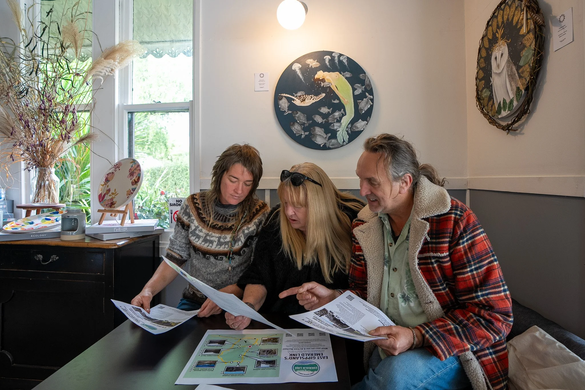 Three people sitting at a table, looking at papers, engaged in discussion, inside a well-lit room with paintings on the wall.