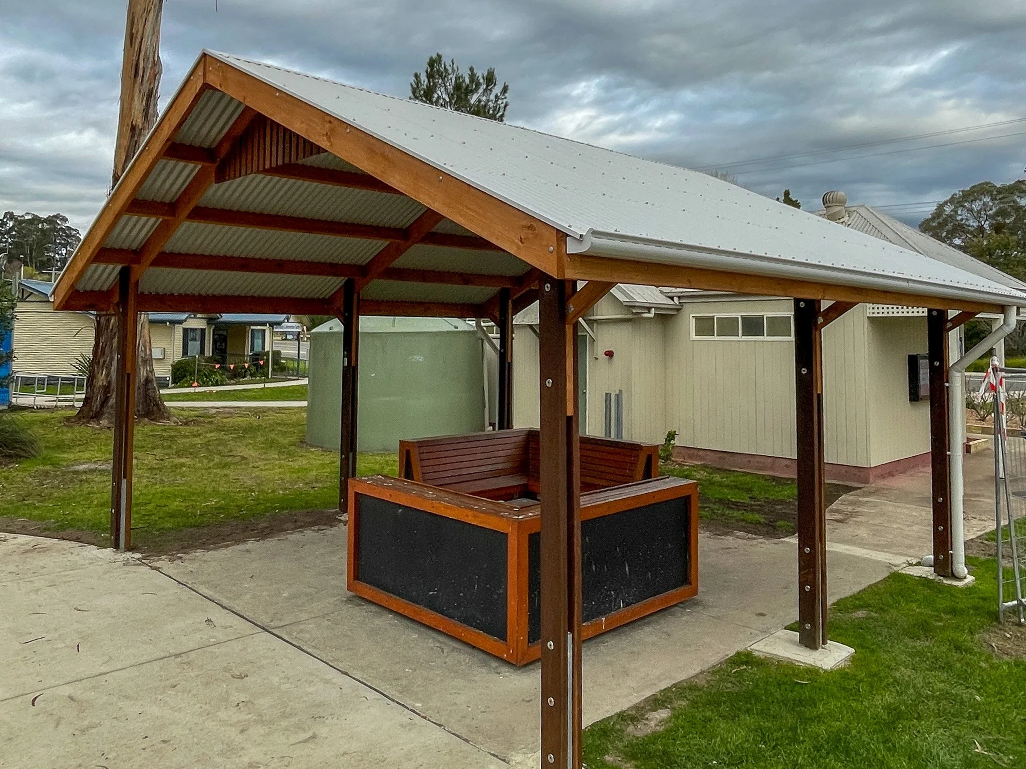A wooden outdoor gazebo with a metal roof and a built-in bench, situated on a concrete pad next to a grassy area and a light-colored building, under a cloudy sky.
