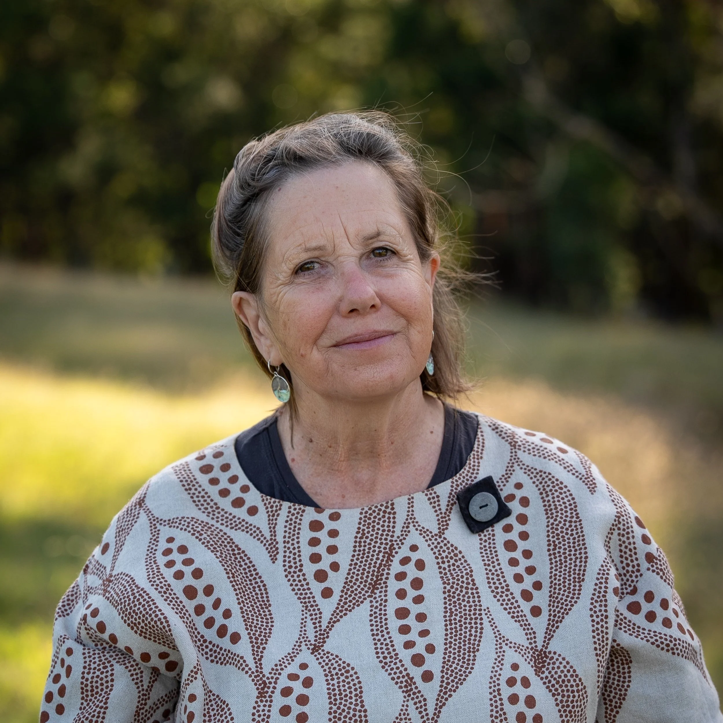 An elderly woman outdoors with a gentle smile, wearing a patterned cream and black top and turquoise earrings, set against a background of trees and greenery.