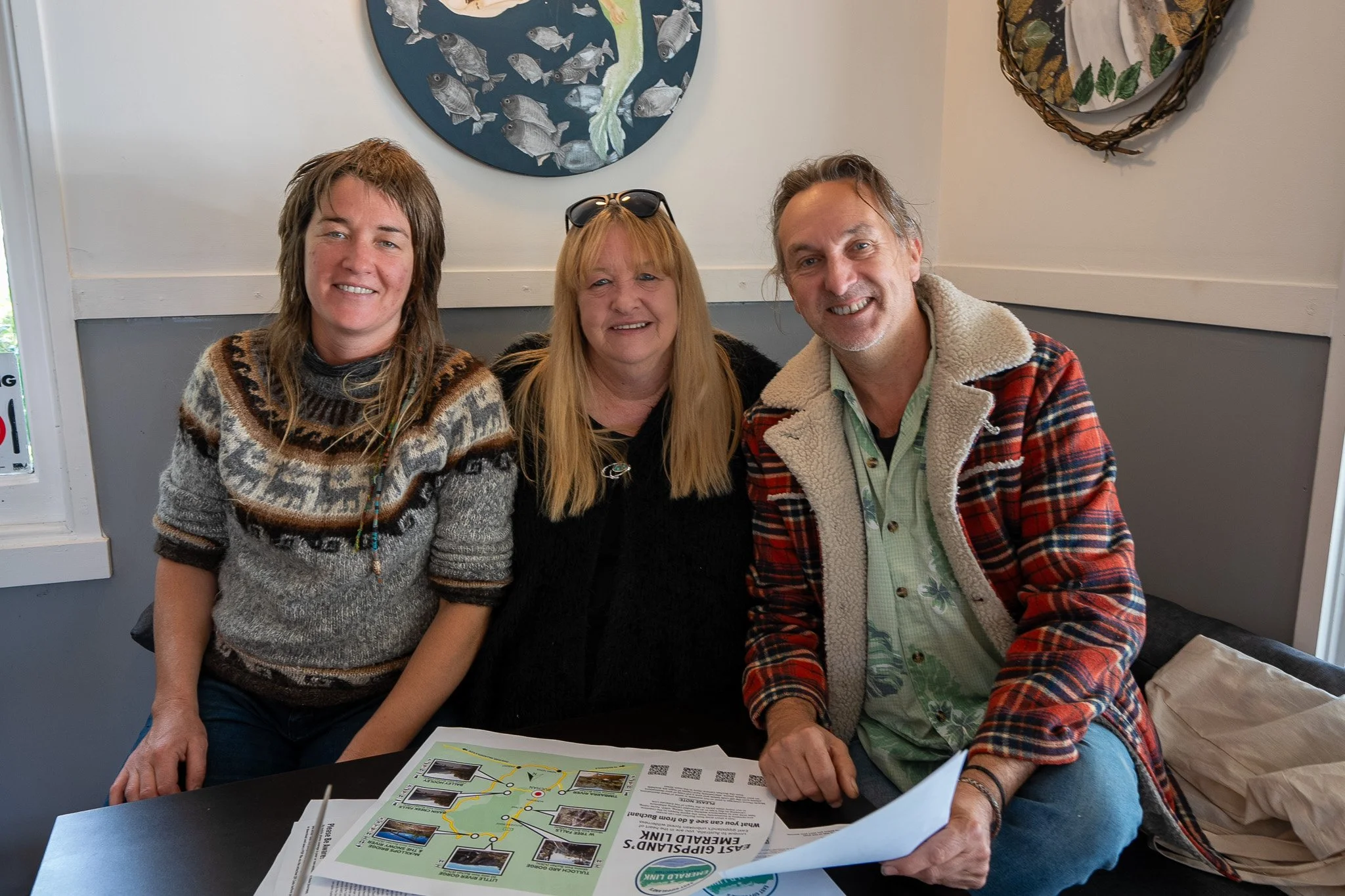 Three smiling people sitting at a table with a map or brochure, in a room with fish-themed artwork on the wall.
