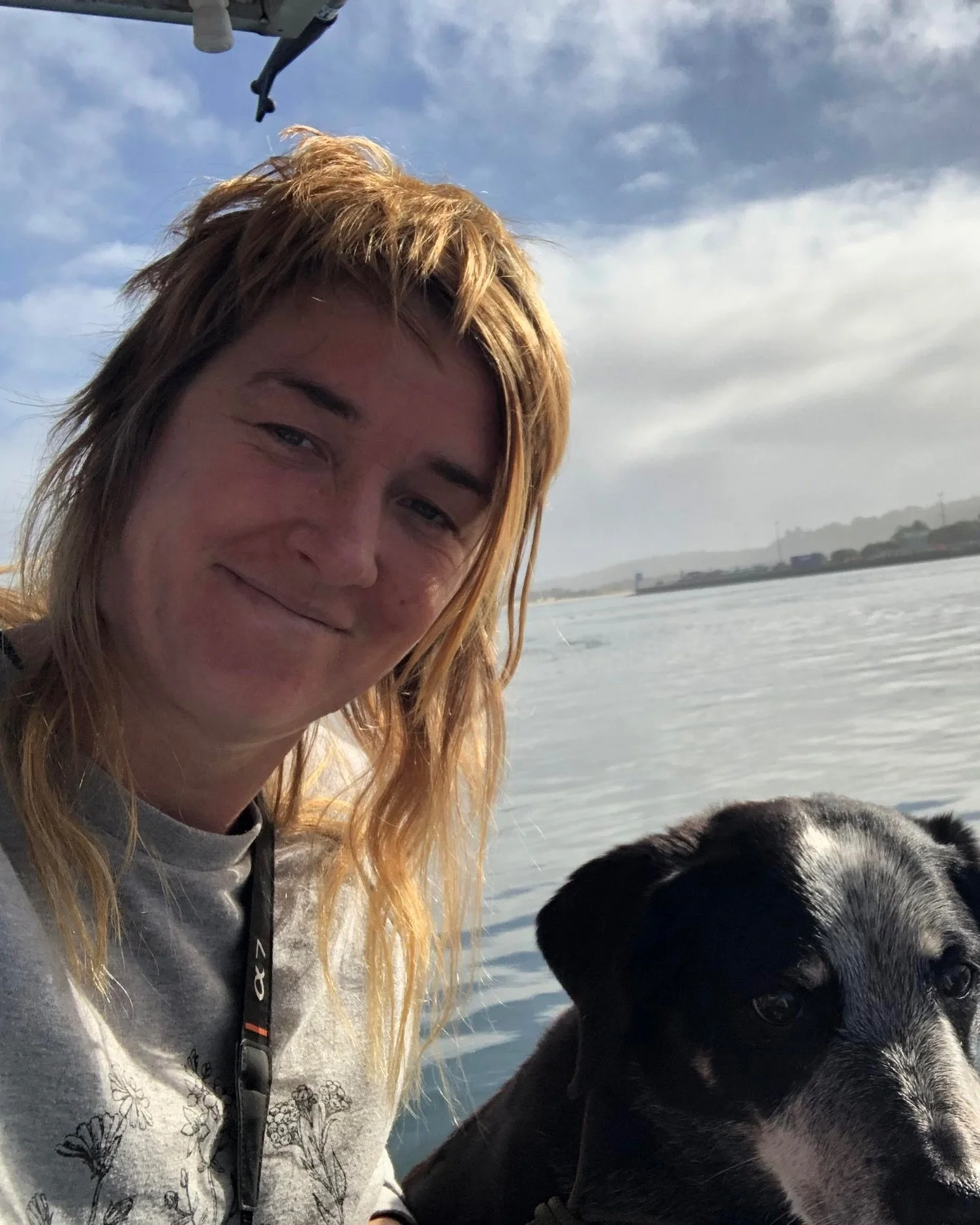 A person with long, blonde hair smiling next to a black and white dog, on a boat with water and cloudy sky in the background.