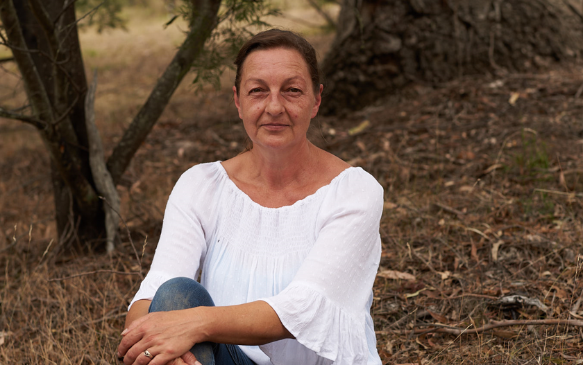 A woman in a white blouse sitting outdoors on the ground with trees and dry grass in the background.