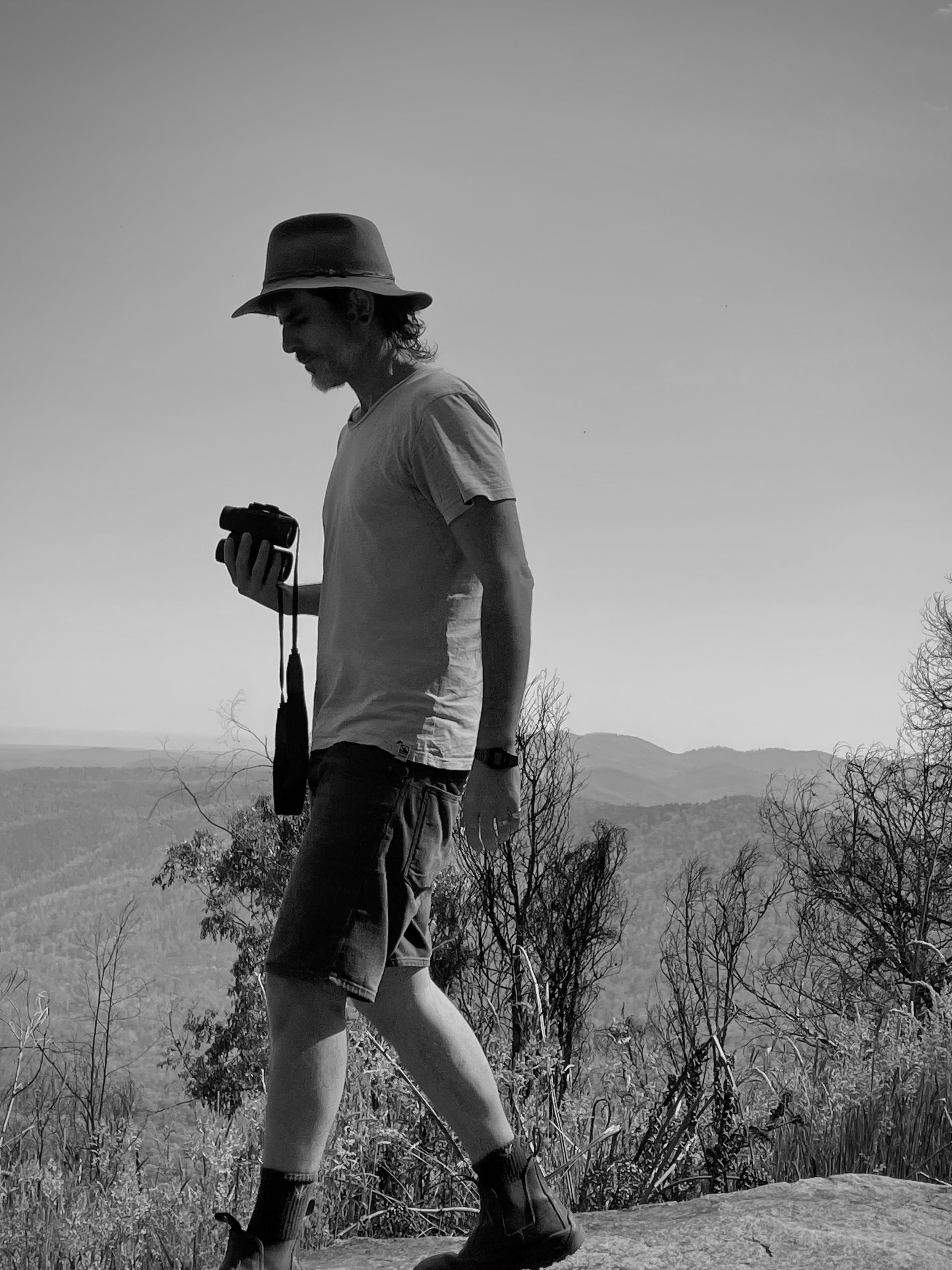 Silhouette of a man walking on a trail at a mountain viewpoint, looking at a camera, with trees and distant mountains in the background, black and white photo.