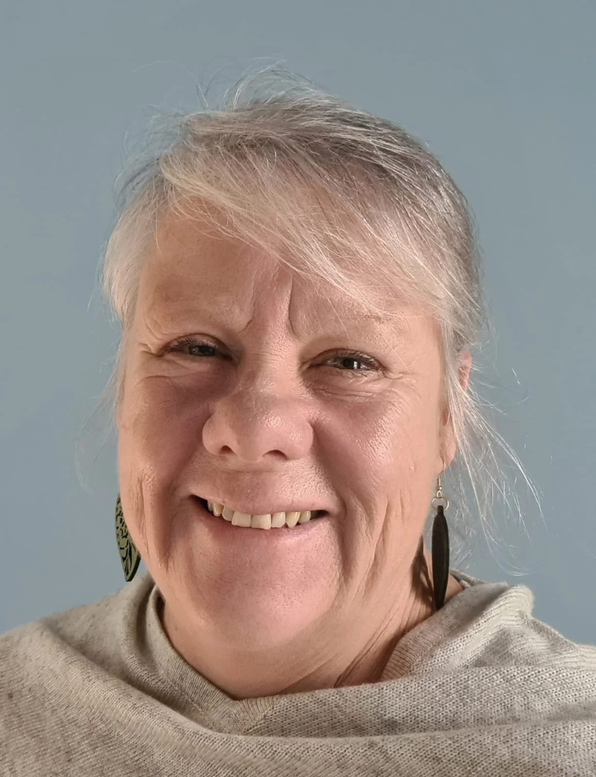Close-up of a smiling older woman with short, silver hair, wearing feather earrings, and a light-colored shawl against a plain background.