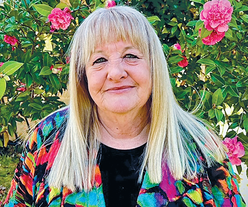 A smiling elderly woman with long blonde hair, standing outdoors in front of pink flowering bushes, wearing a colorful patterned jacket.
