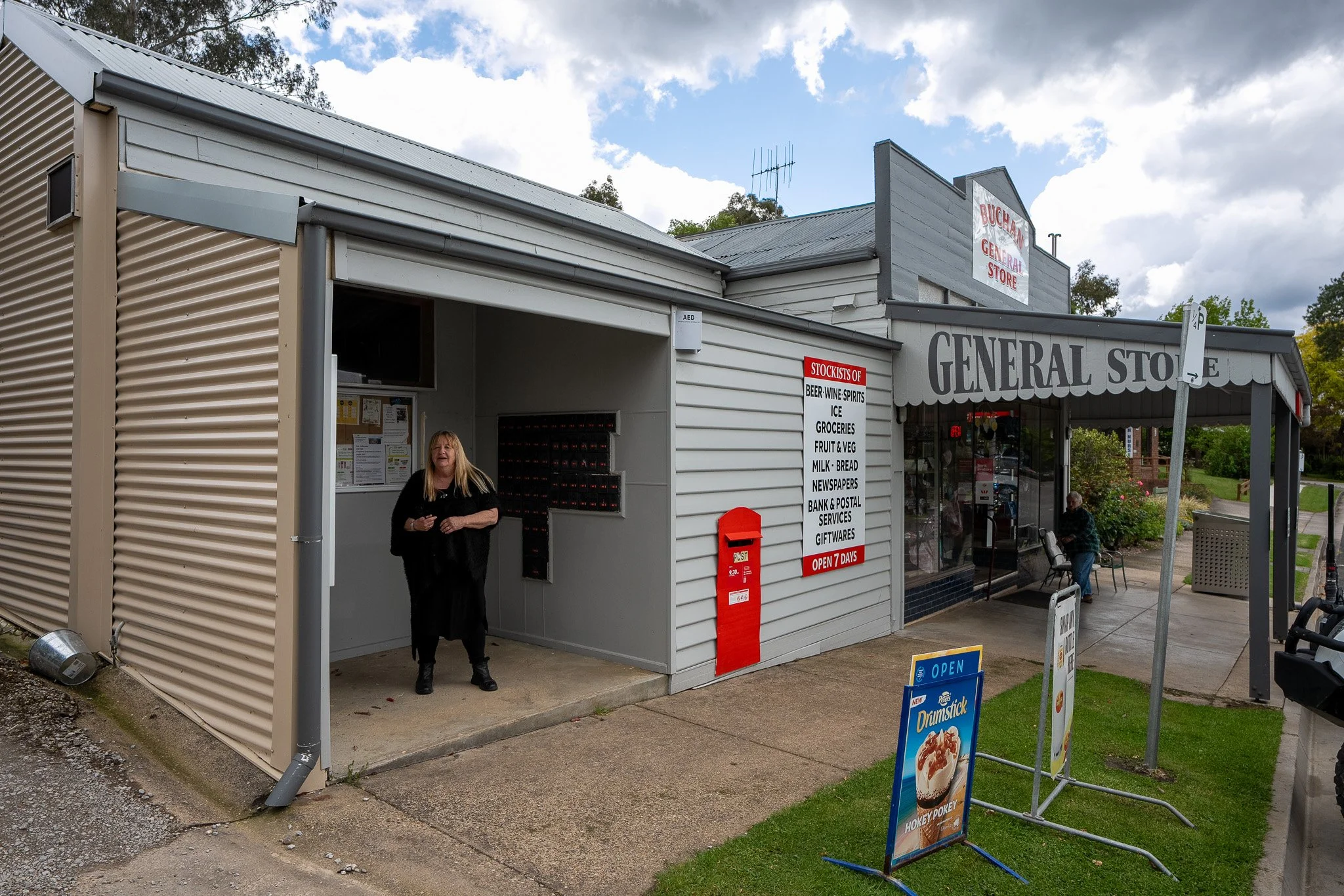 A small general store with a gray and white exterior, a porch with two chairs, and a woman standing near a vending machine. There are signs for stock items, an open sign, and advertisements for food outside. The sky is partly cloudy.