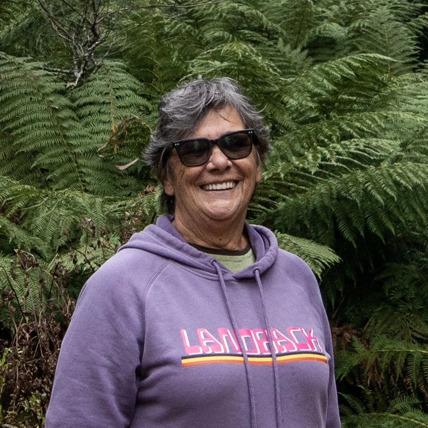 A smiling woman with short gray hair wearing sunglasses and a purple hoodie standing in front of large green fern leaves.