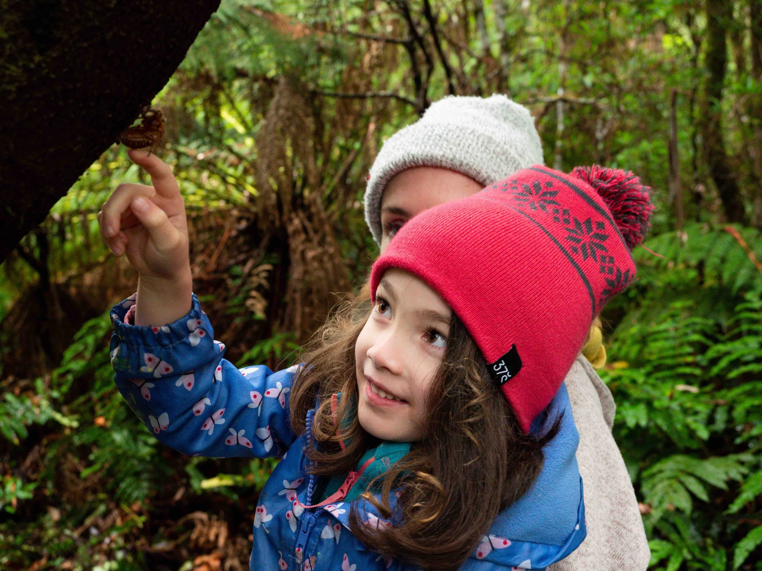 A young girl and woman, wearing warm hats and jackets, are in a forest. The girl is pointing at a small insect on a tree trunk while the woman looks on, surrounded by greenery.