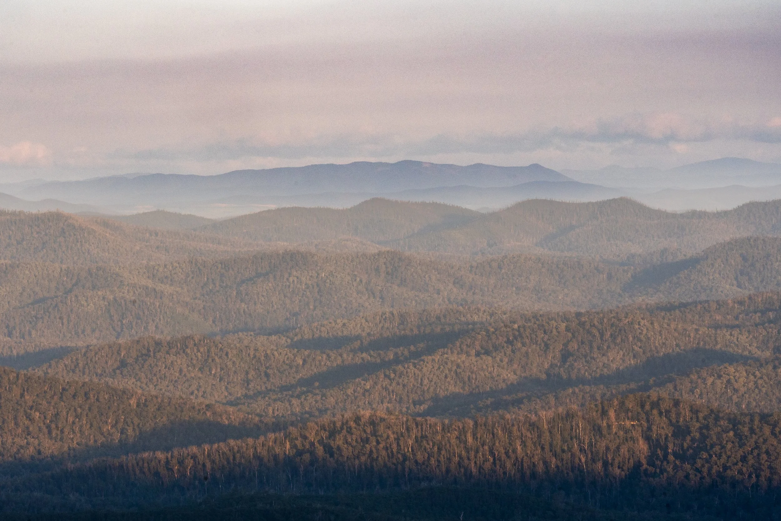 Landscape of rolling forested hills and mountains under a cloudy sky with distant mountain ranges in the background.