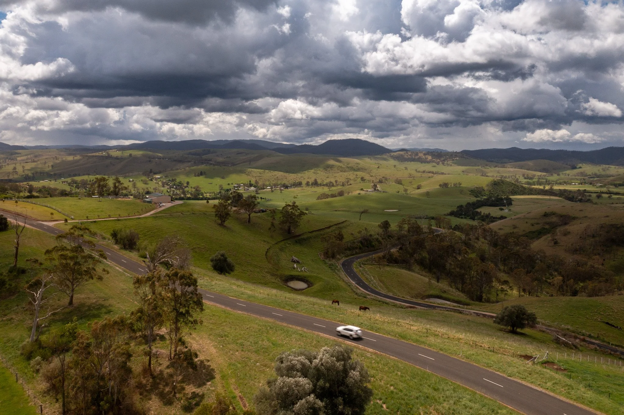 Scenic view of rolling green hills with a winding road, scattered trees, small ponds, and distant mountains under a cloudy sky.