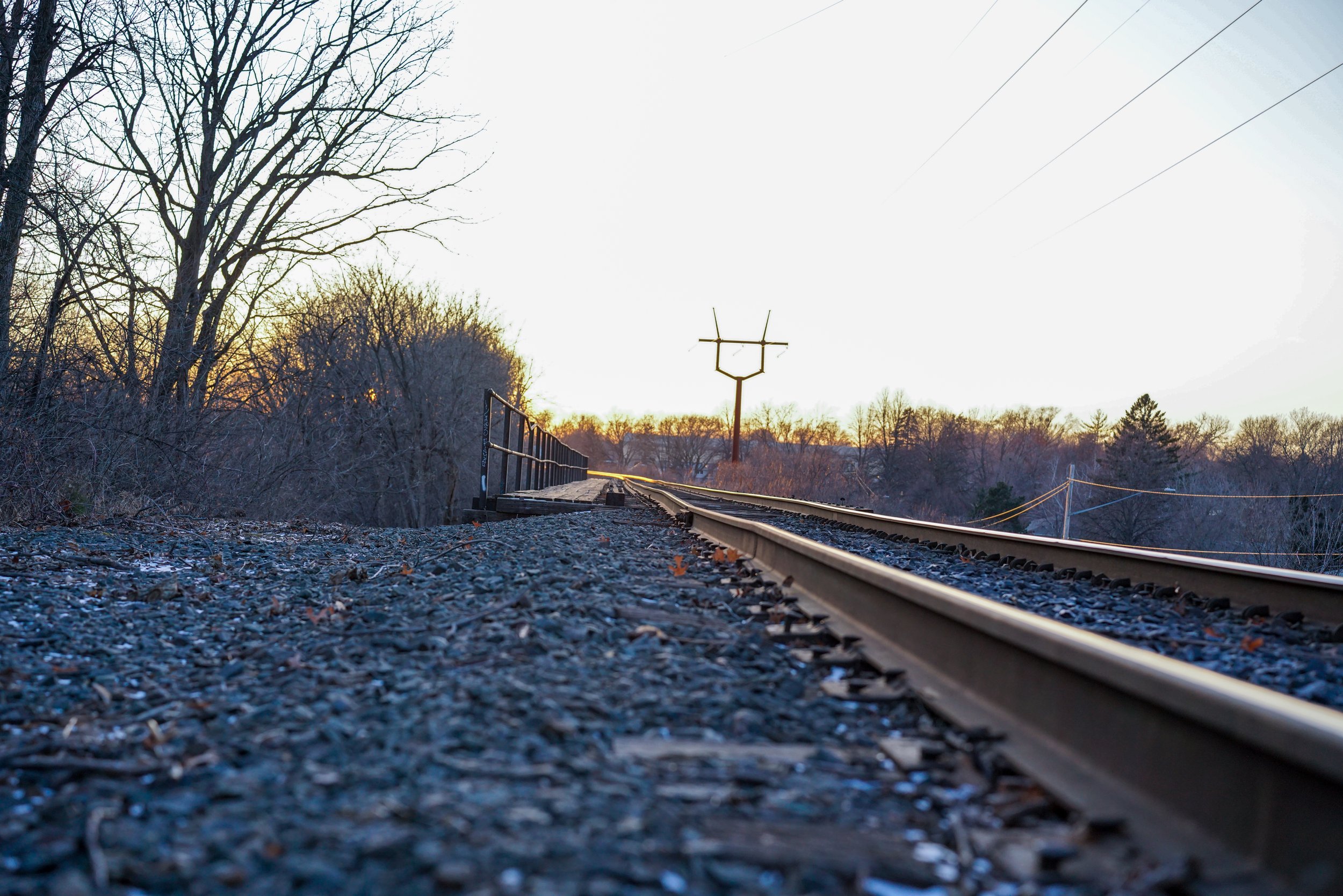 A view of a railroad track extending into the distance during sunset, with leafless trees on the left and utility poles on the right.