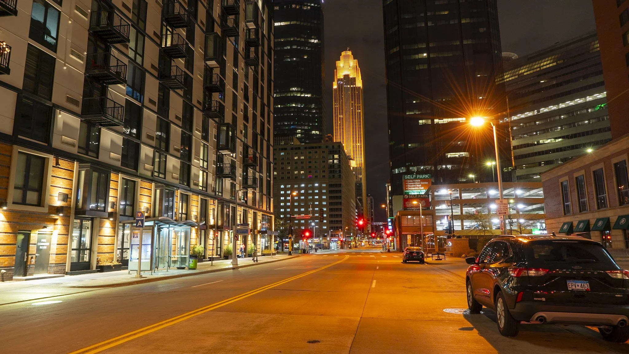 Nighttime city street scene with modern buildings, illuminated windows, streetlights, and a tall skyscraper with golden lighting in the background.