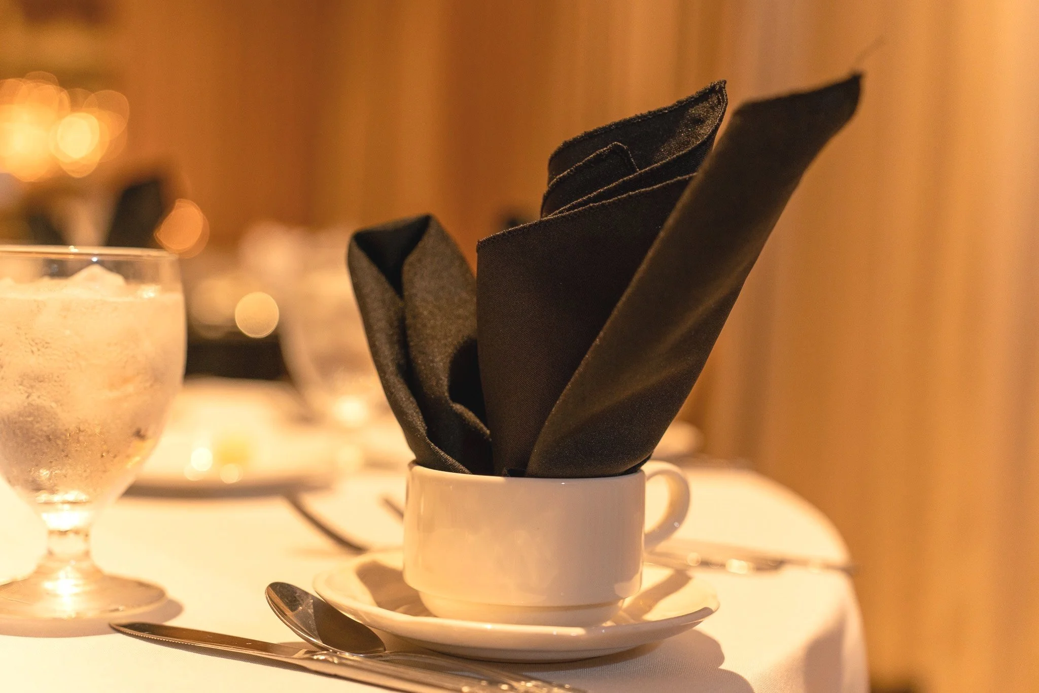 A white cup with black napkins folded inside, placed on a small white saucer with a metal spoon, on a table set for dining with a lit candle in the background.