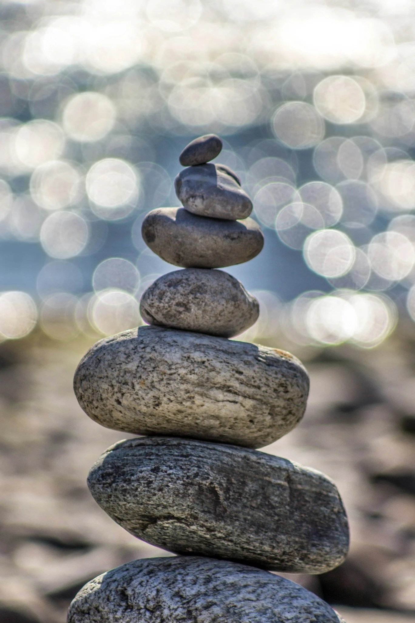 A balanced stack of seven smooth, rounded rocks on a beach with blurred water and sunlight reflections in the background.